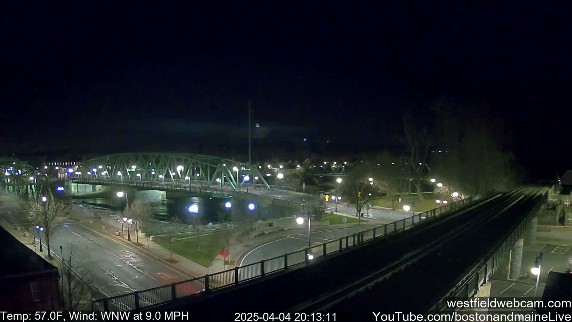A nighttime view of a bridge over a river, with a road and train tracks intersecting nearby under a clear sky, and a temperature of 57°F with a west-northwest wind at 9.0 mph.