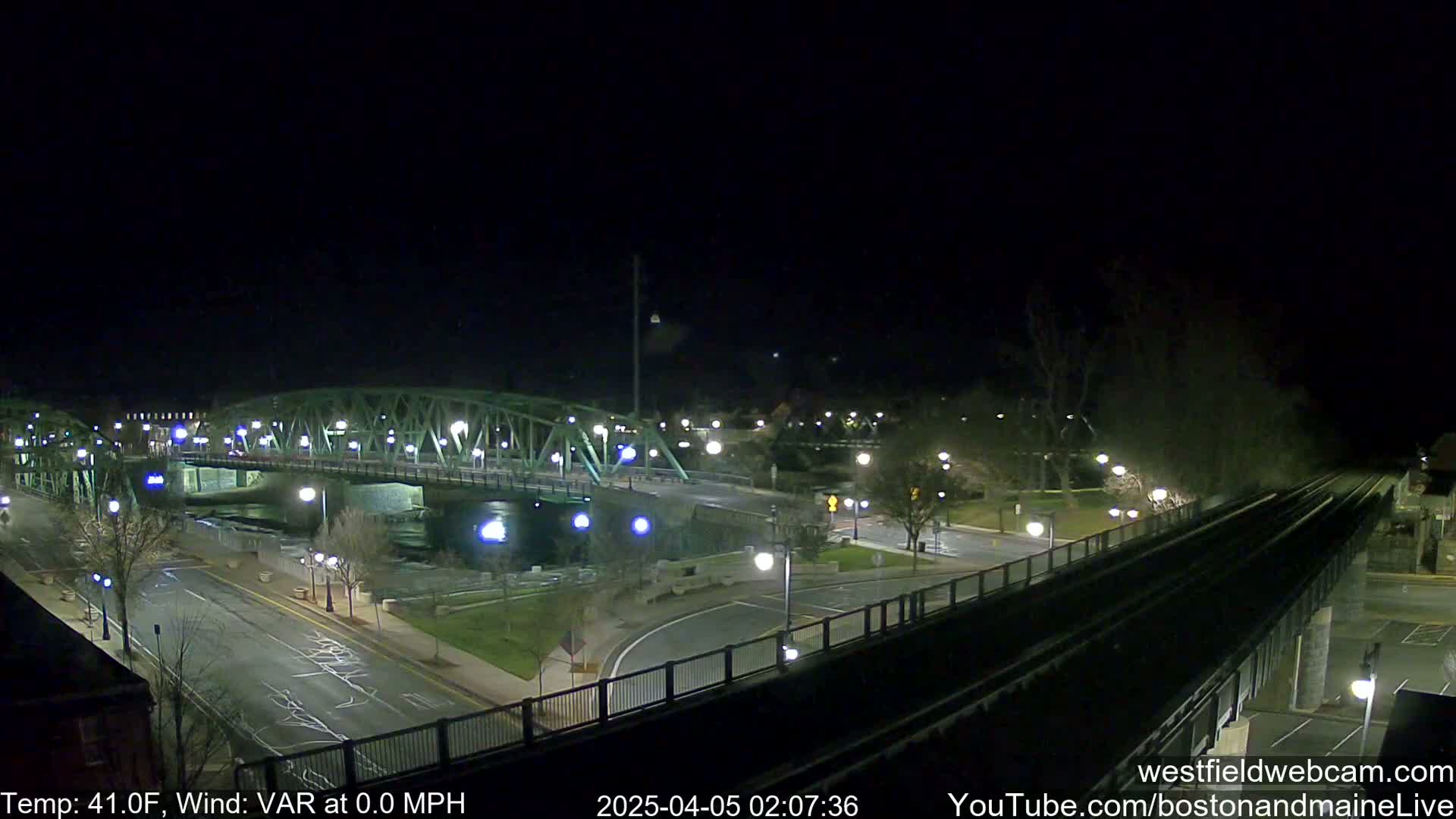 A nighttime view of a bridge over a river, with a road and train tracks visible, under clear skies.