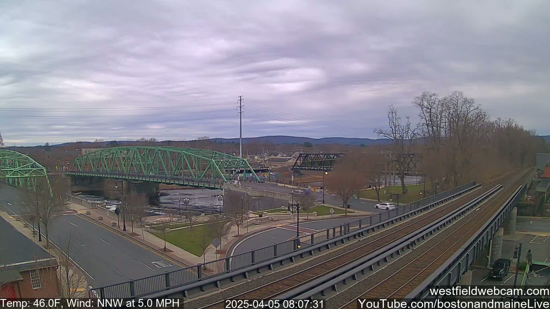 A view of two bridges over a river, one green and one black, with train tracks and a road in the foreground, under a cloudy sky.