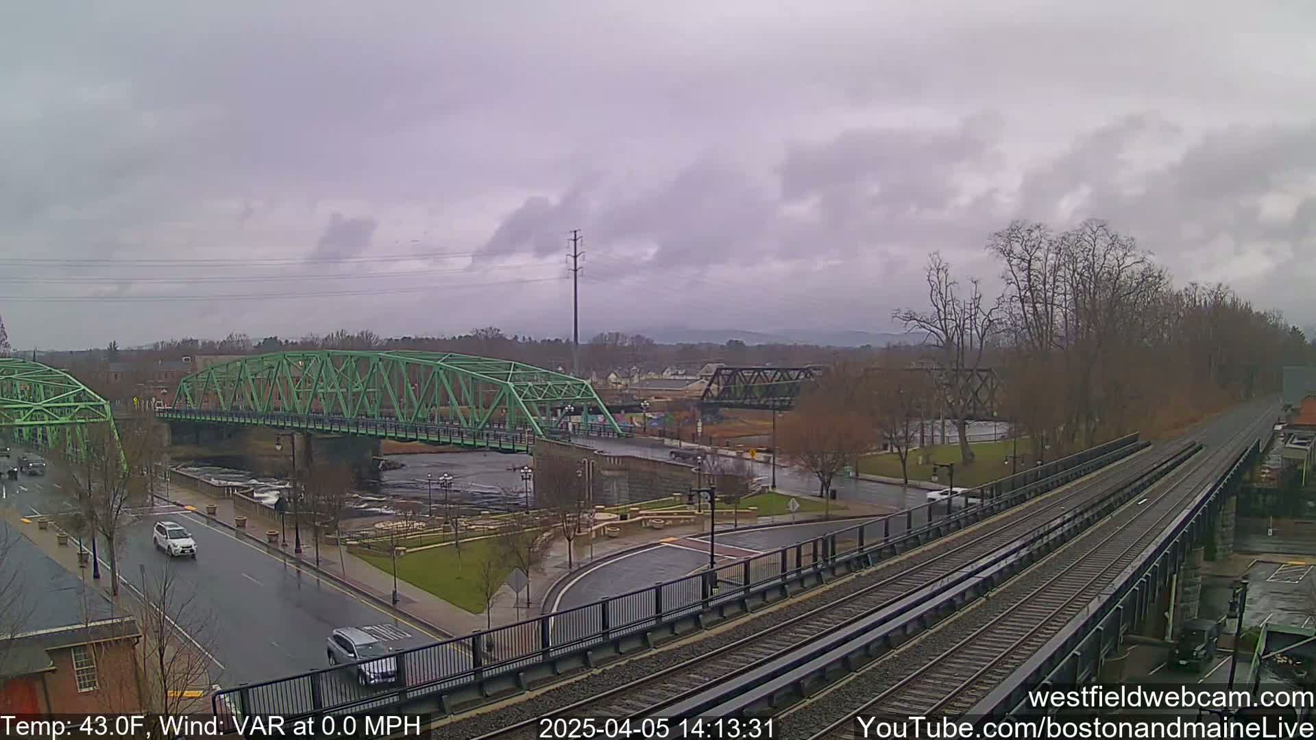 A green bridge spans a river under an overcast sky, with train tracks and cars visible on a nearby road.