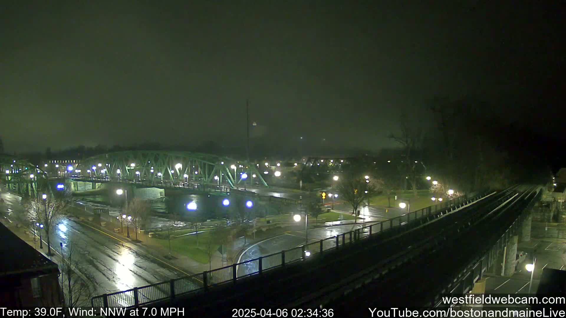 A nighttime view of a rain-slicked bridge and roadway, with a railroad track in the foreground, under a dark, overcast sky.