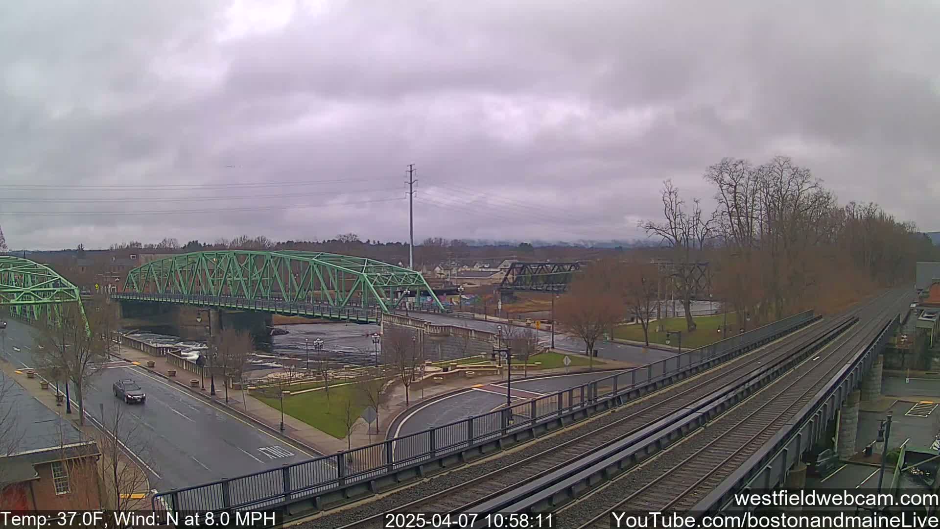 A green bridge spans a river under an overcast sky, with train tracks and a road visible in the foreground.