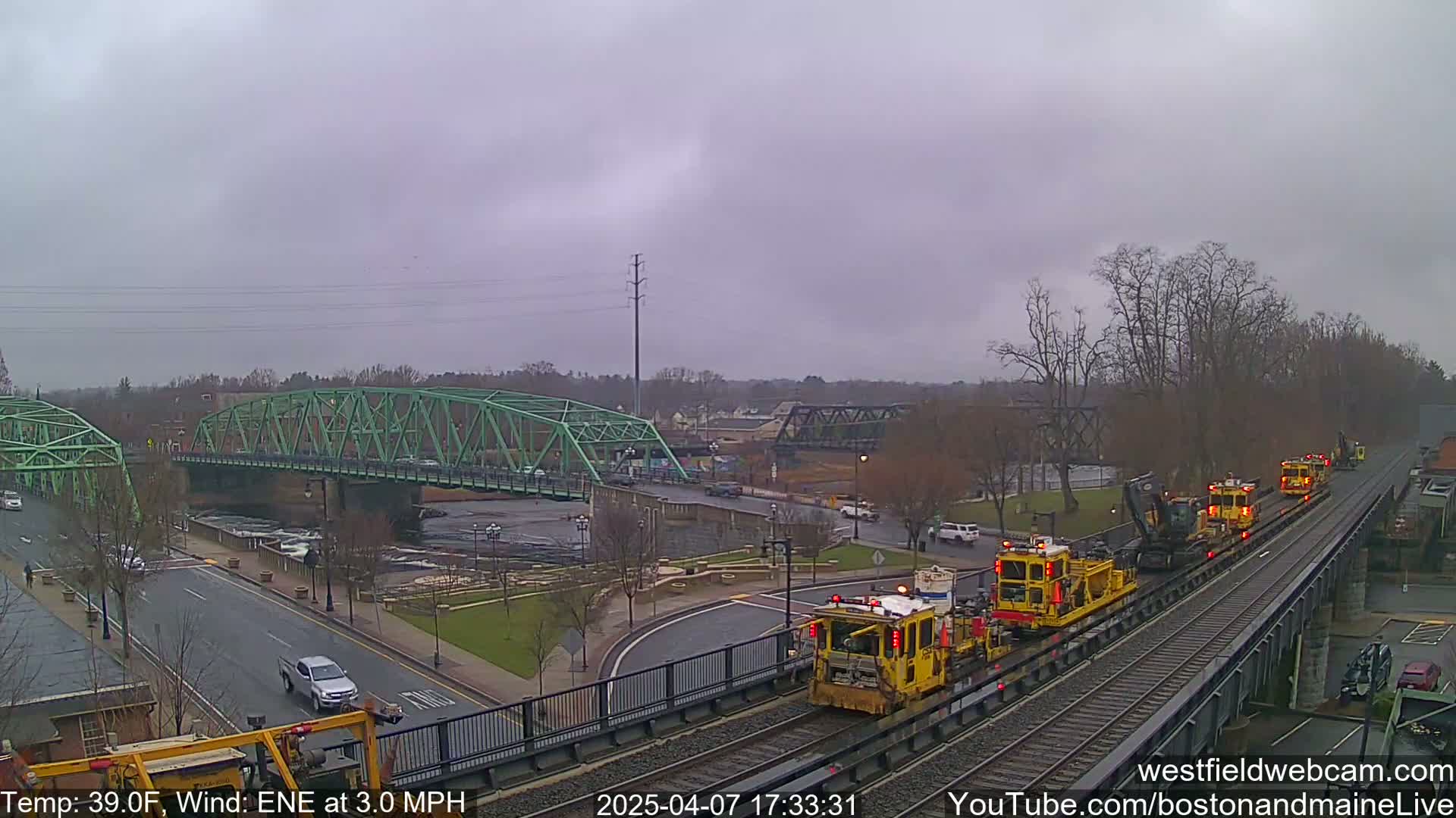 A train of yellow maintenance vehicles travels on railroad tracks alongside a river, under overcast skies, passing near a green bridge.