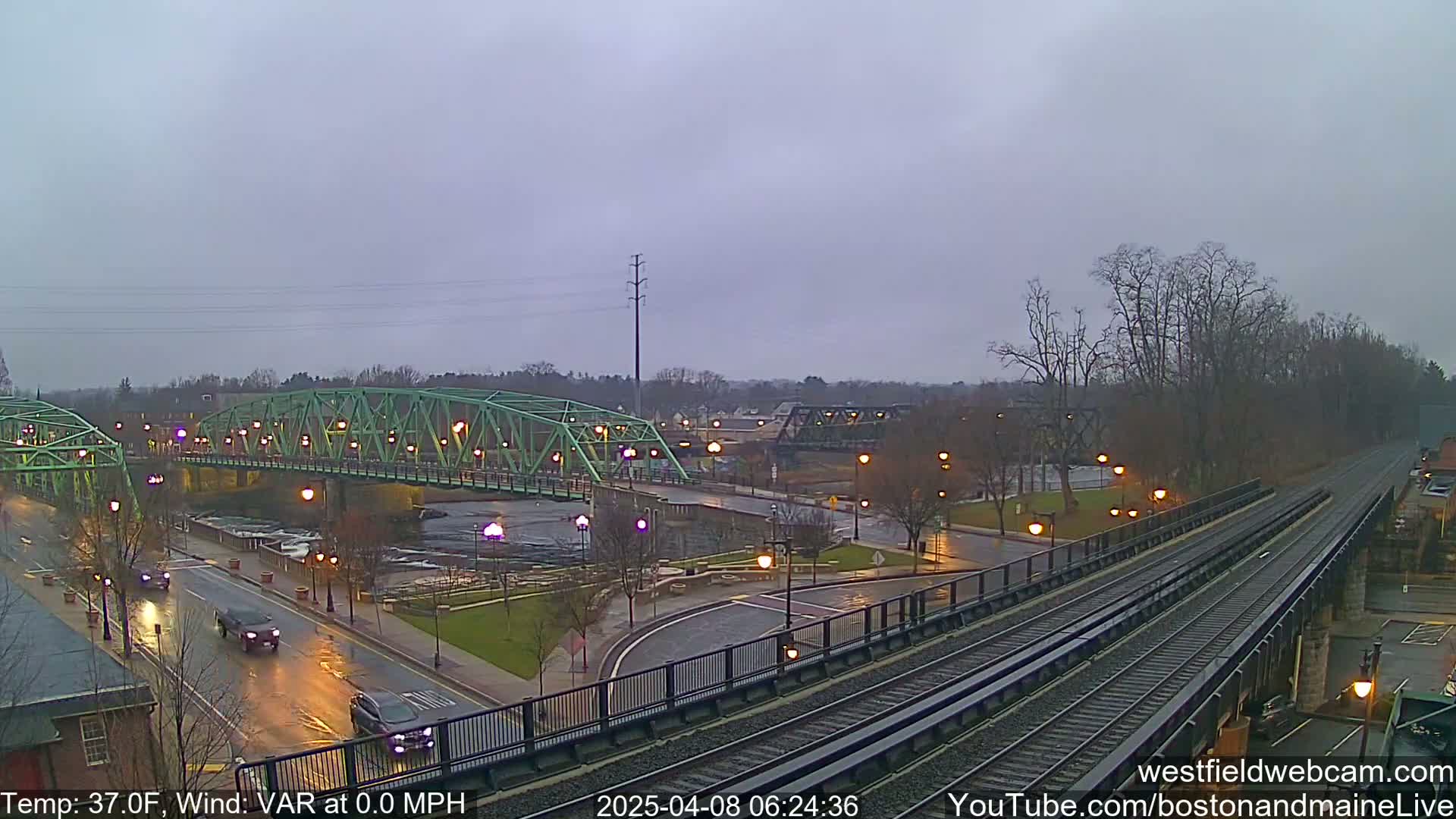 A wet, overcast scene shows a green bridge over a river, a roadway with cars, and train tracks leading away from the viewer.