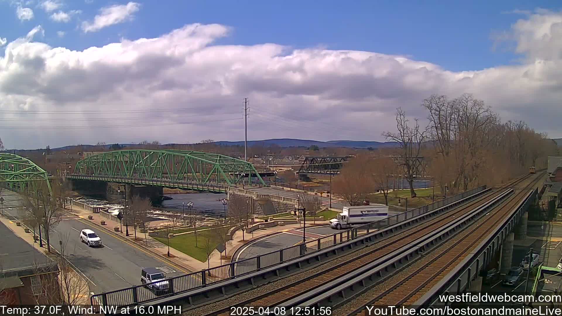 A partly cloudy day with a northwest wind at 16 mph shows a view of two bridges spanning a river, a roadway with vehicles, and railroad tracks running alongside.