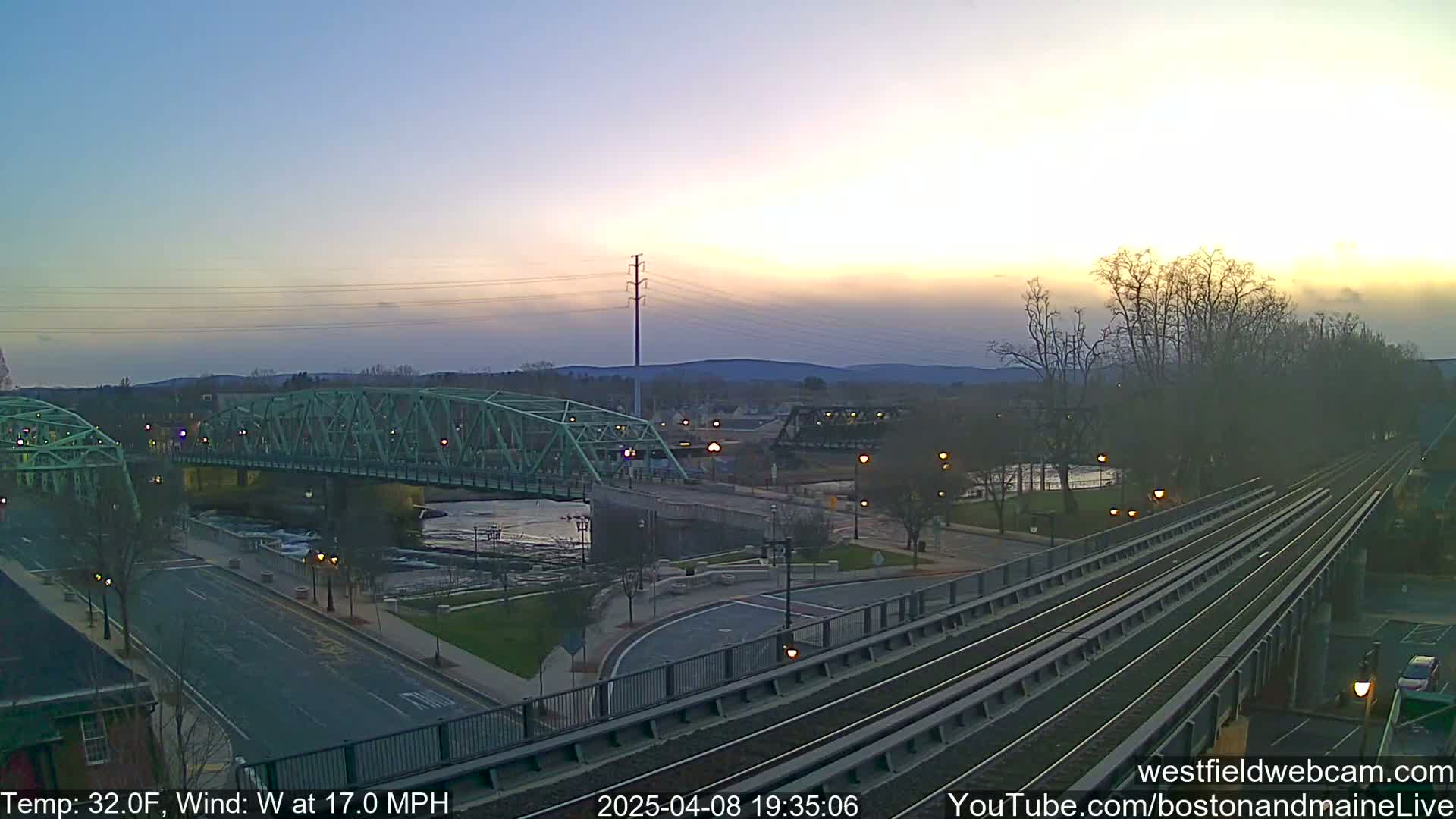 A light-green bridge spans a river at dusk under a mostly clear sky, with train tracks and a road visible in the foreground, and a temperature of 32°F and a wind of 17.0 MPH.