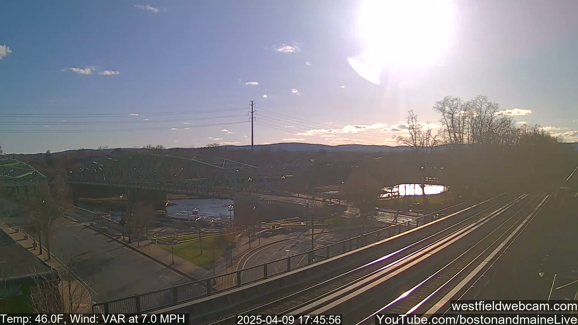 A high-angle view of a river, bridge, train tracks, and a town under a partly sunny sky with a 7.0 mph wind.