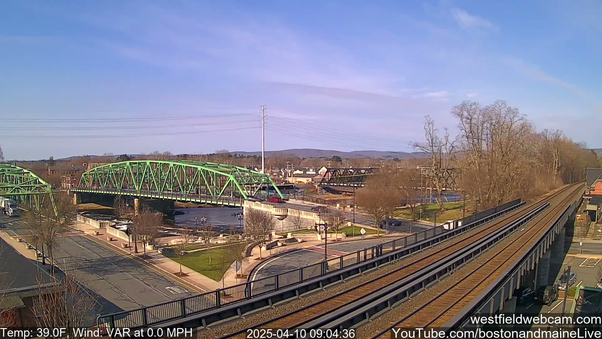 A view from an elevated train track shows a green bridge spanning a river in a town under a clear, sunny sky.