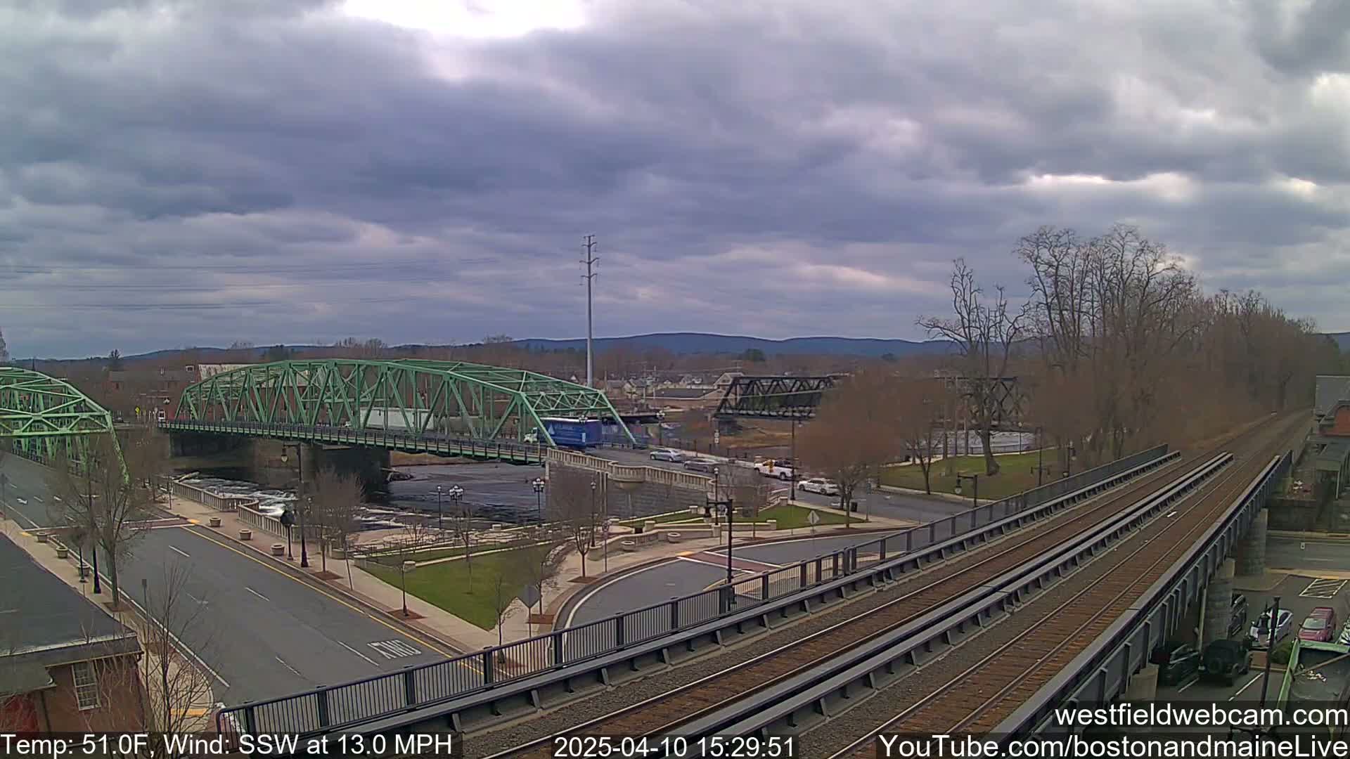 A view of a town with two bridges over a river, train tracks, and a partly cloudy sky, with a wind blowing from the south-southwest at 13 mph.