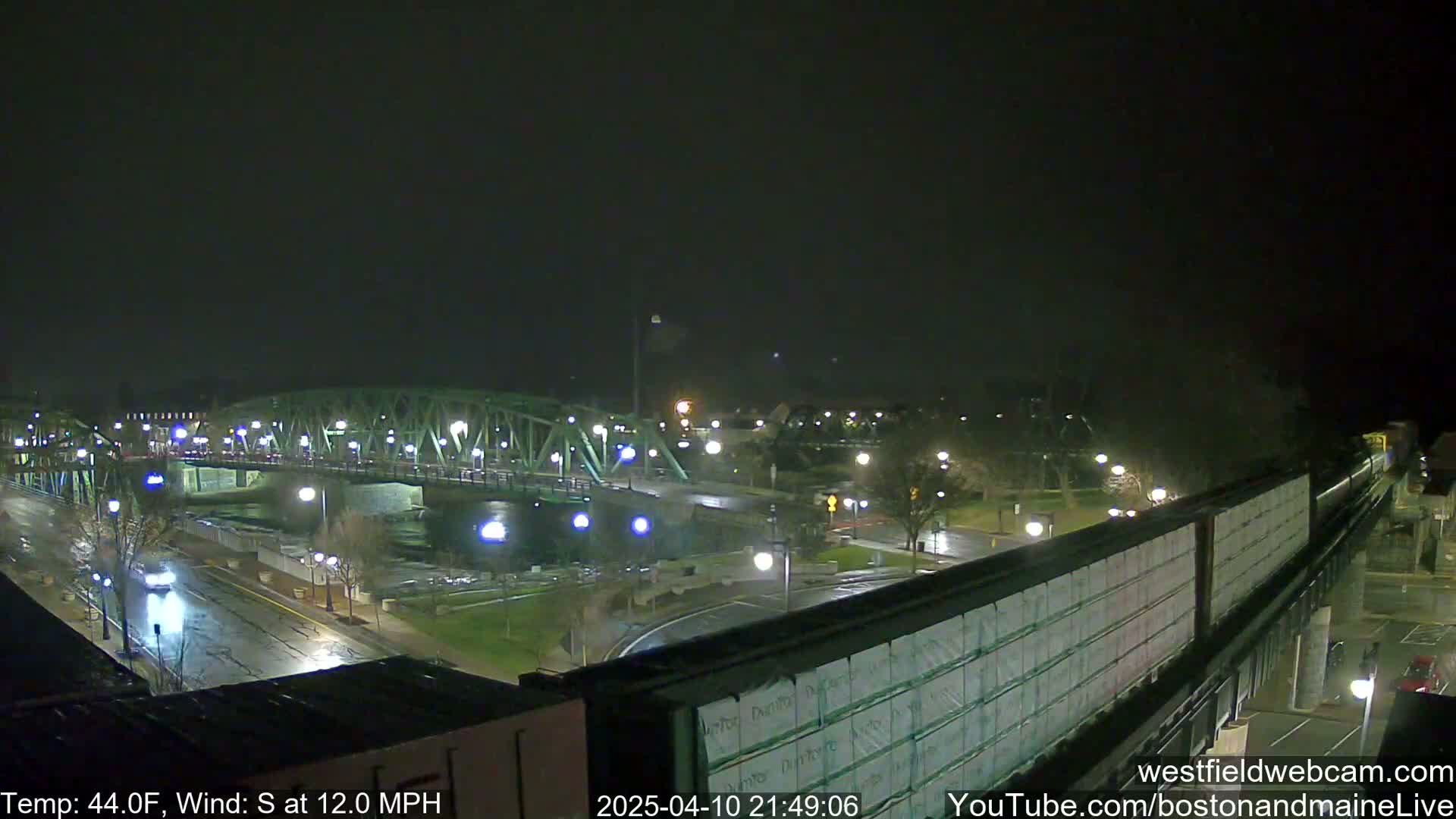 A nighttime view of a bridge over a river, with wet streets and a partly cloudy sky, under a 44°F temperature and 12 mph south wind.