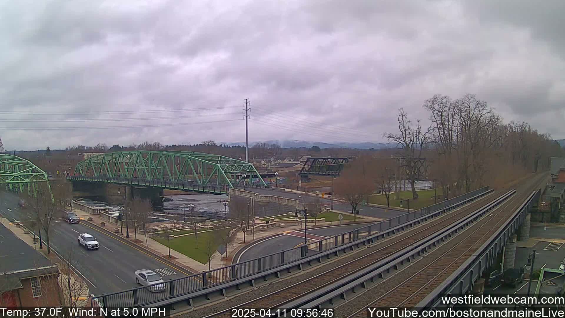 A view of two bridges over a river, with a railway line in the foreground, under an overcast sky, with a gentle northerly wind.