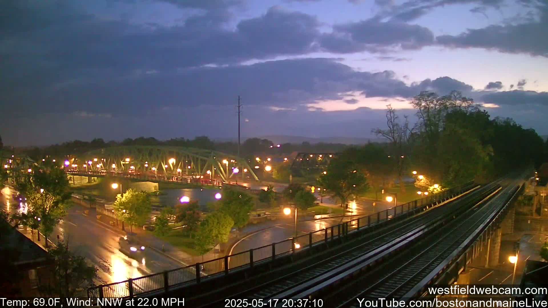 A rain-slicked nighttime scene shows a lit bridge over a river, a roadway with cars, and train tracks, all under a dark, cloudy sky.