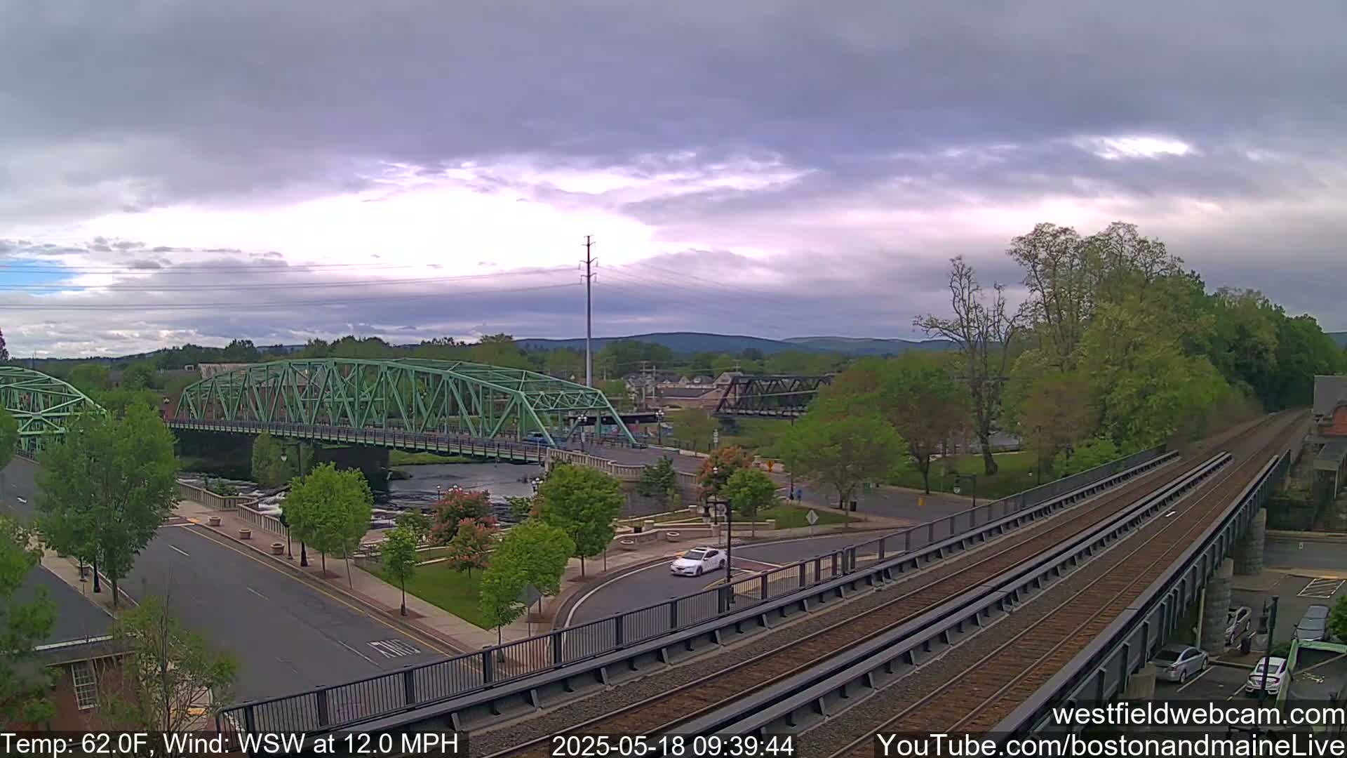 A partly cloudy sky overlooks a town with two bridges, a river, and train tracks, with a light breeze blowing from the west-southwest at 12 miles per hour.