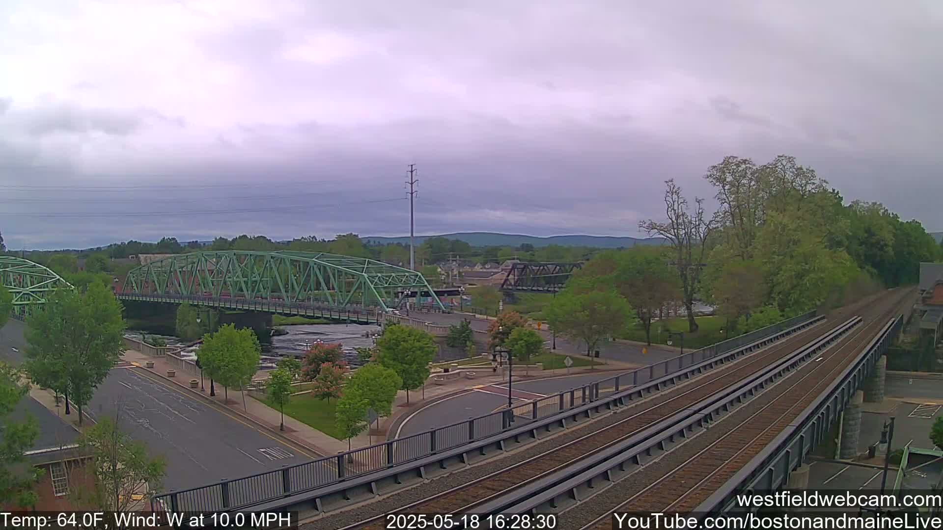 A view of a town on an overcast day shows two bridges over a river, a railway track, and green trees.