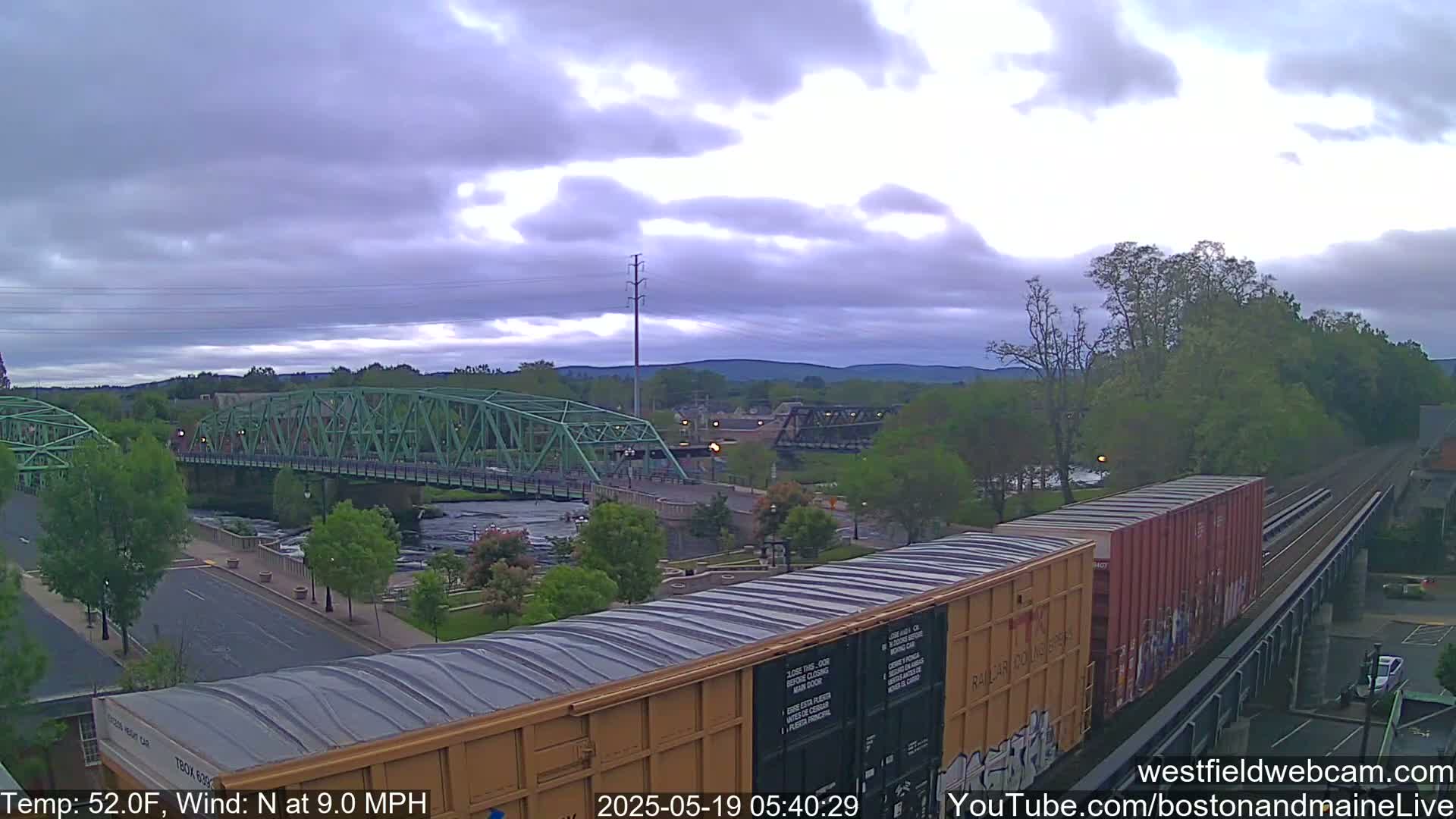 A green bridge spans a river under a cloudy sky, with a train car partially visible in the foreground.