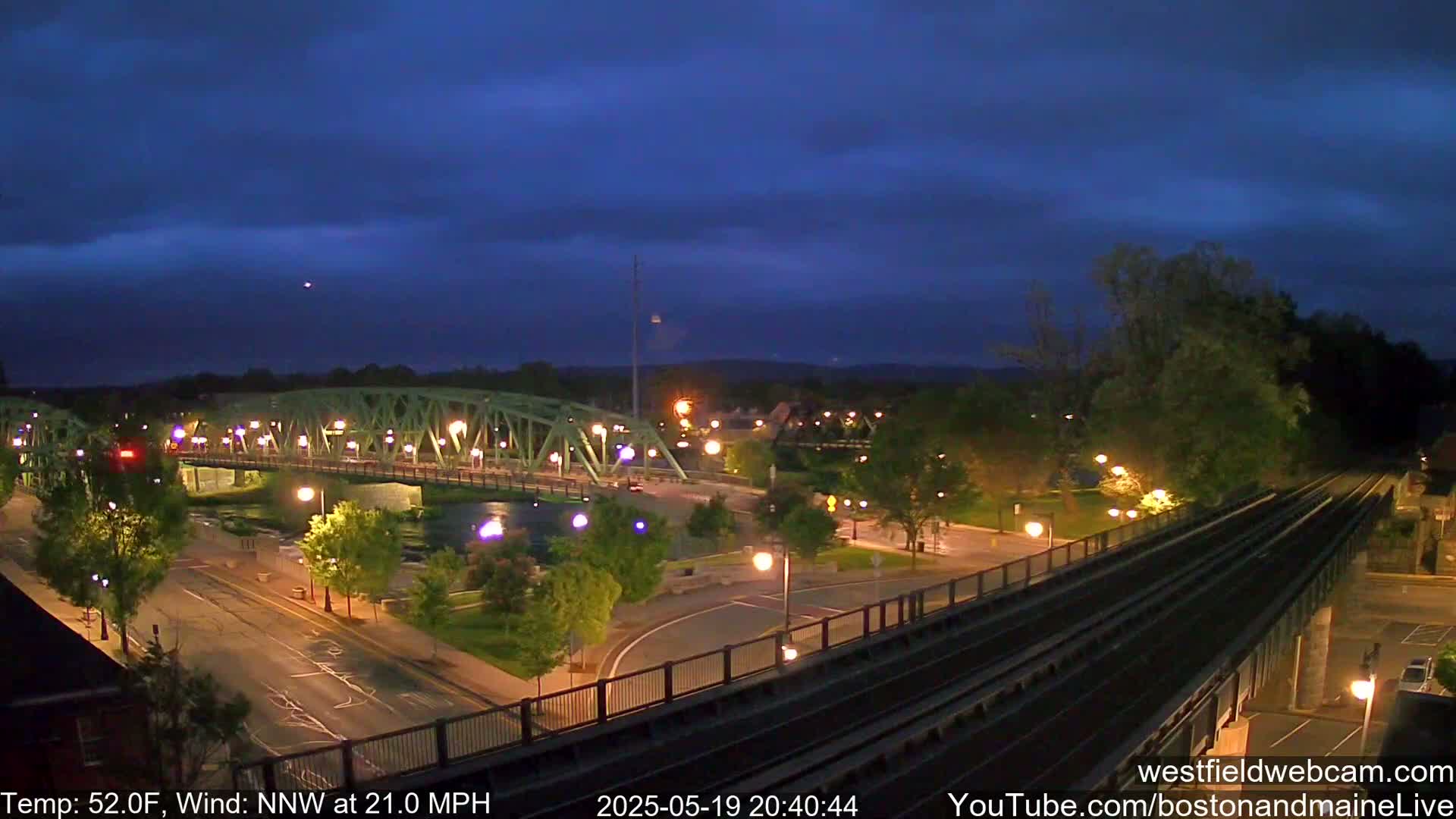 A nighttime view of a bridge over a river, a road, and a set of train tracks under a mostly clear, dark blue sky with a 21 mph northwest wind and a temperature of 52°F.