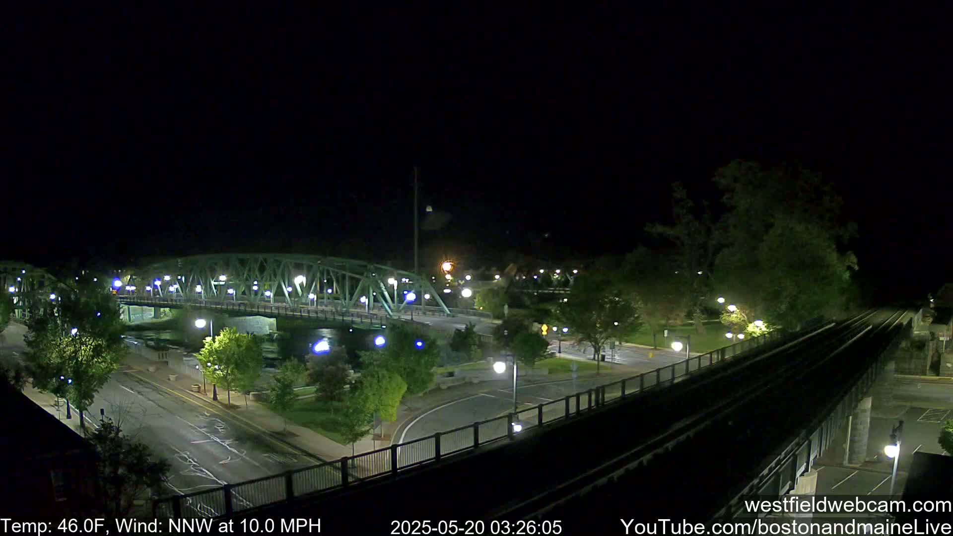 A nighttime view of a bridge over a river, with a roadway and train tracks visible, under a clear sky and a 10 mph north-northwest wind.