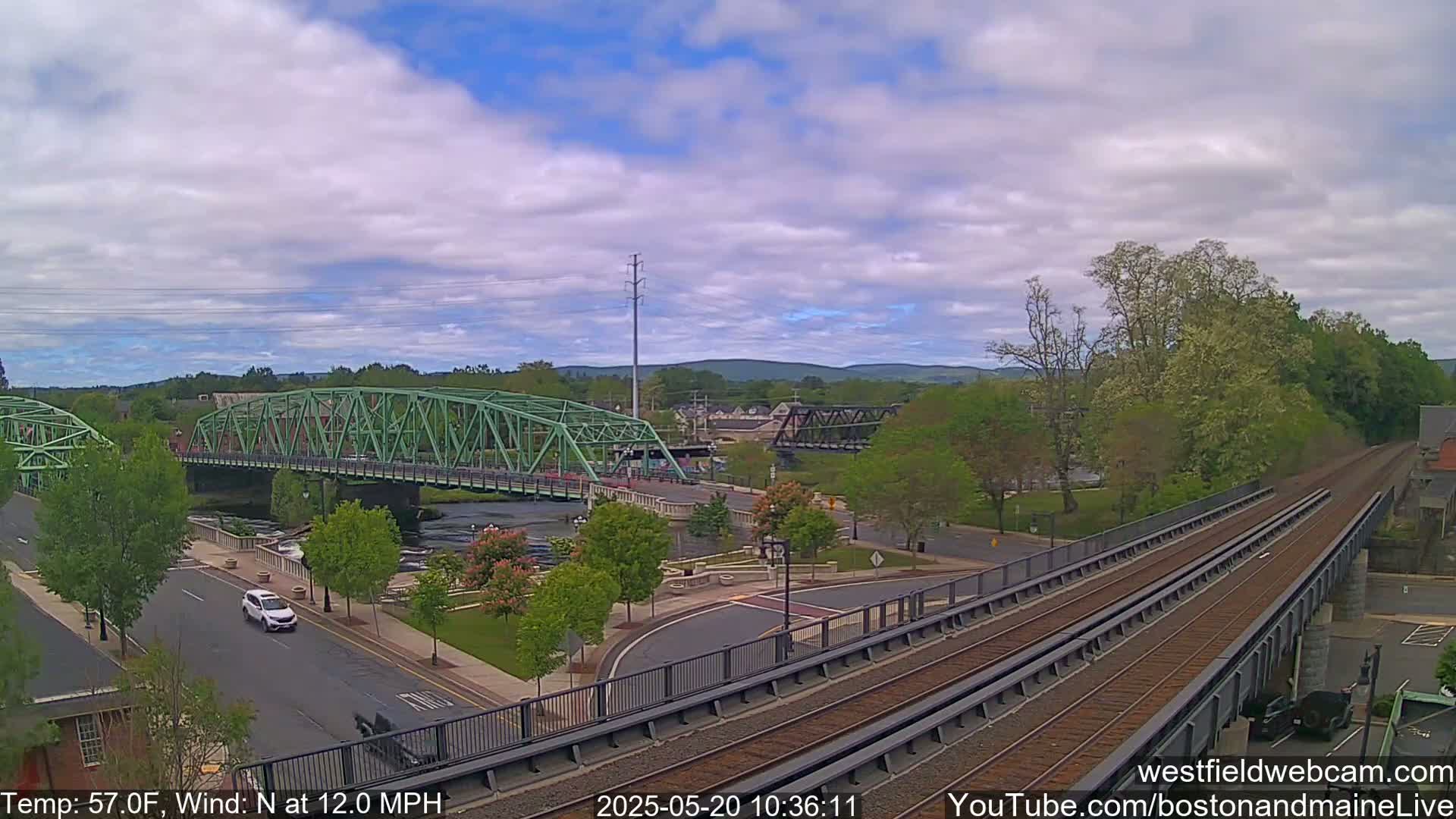 A partly cloudy day shows two bridges over a river, with train tracks and a roadway intersecting in a town setting.