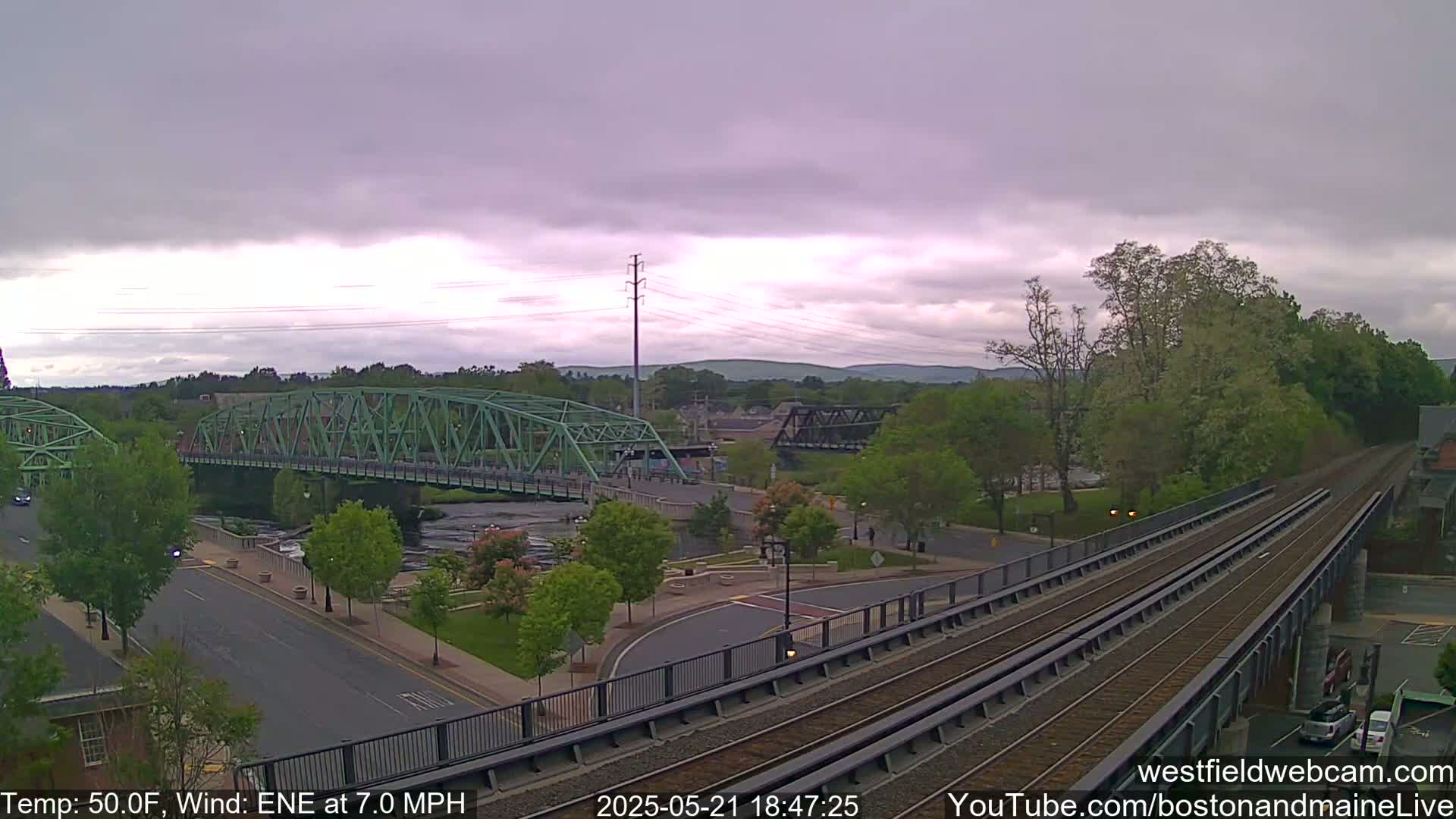 A view of a river with two bridges, one green and one dark metal, under a cloudy sky, with train tracks in the foreground and leafy green trees and roads surrounding the river.