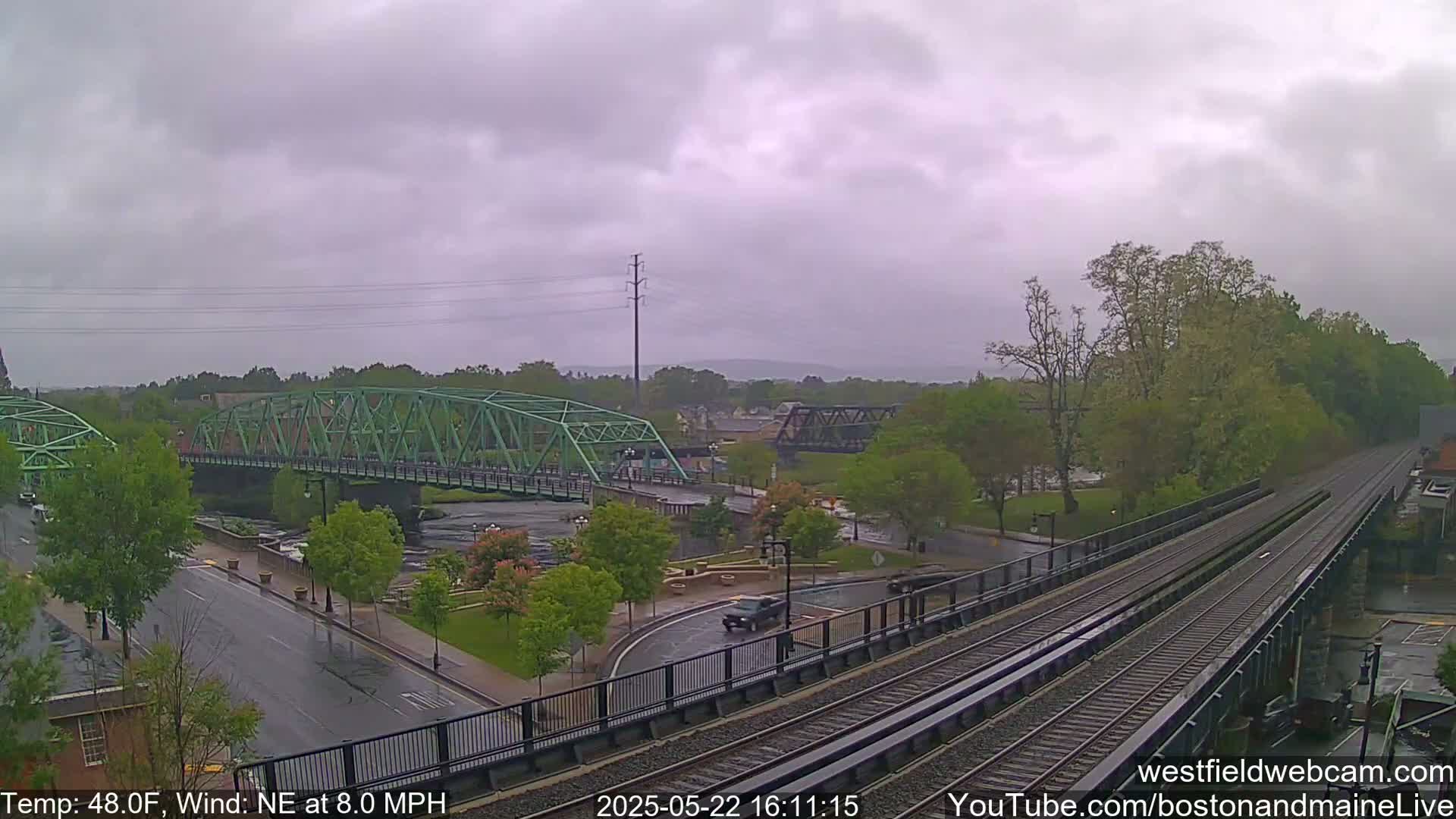 A view of a town on an overcast day with rain, showing two bridges over a river, a roadway, and train tracks.