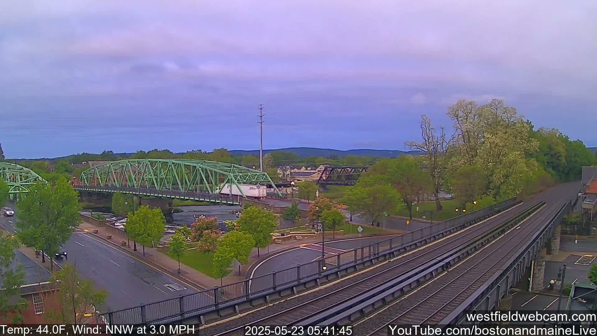 A green metal bridge spans a river, with train tracks and a roadway alongside it, under a mostly clear, light-purple sky.