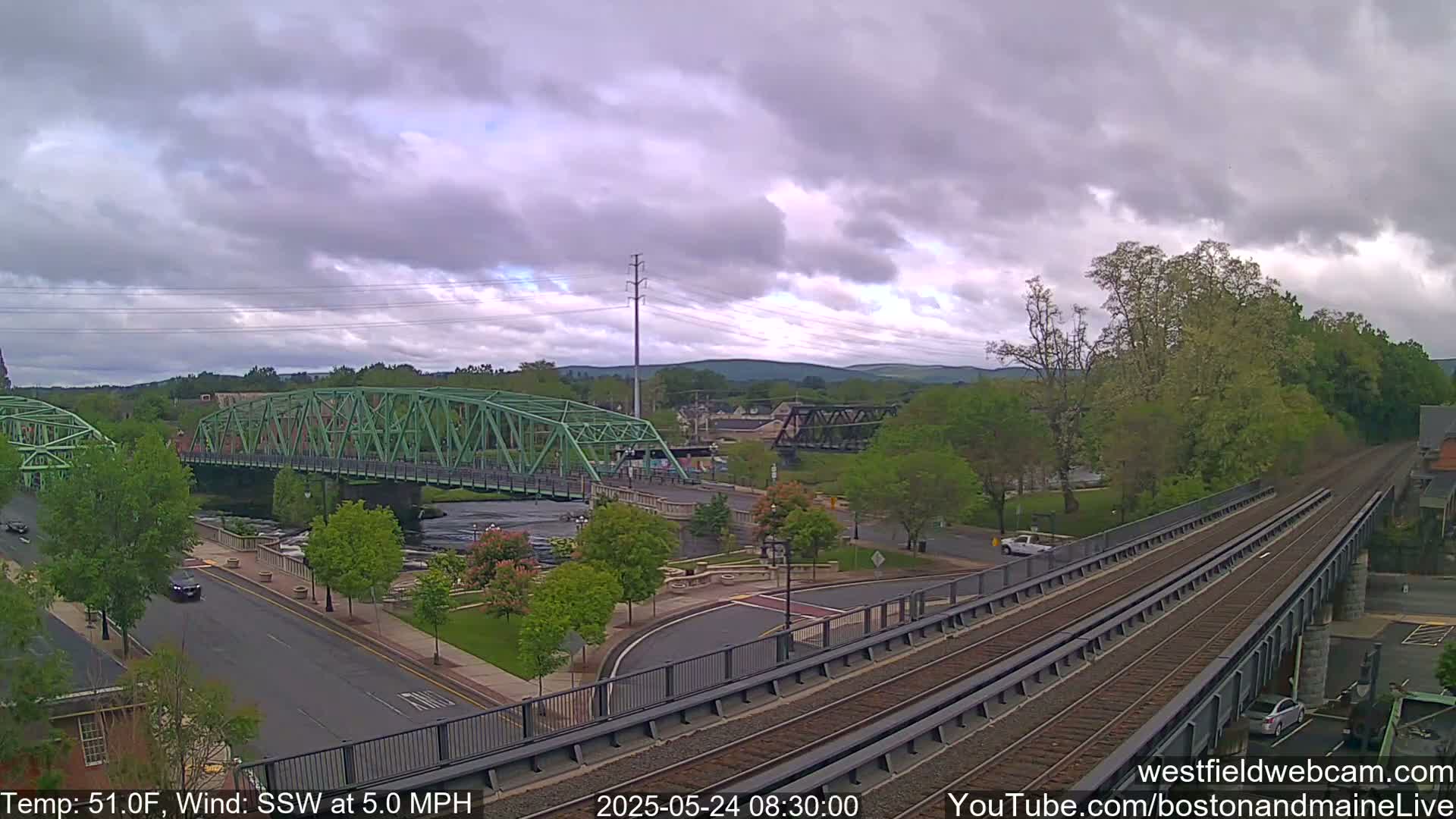 A view of a town on an overcast day, showing a green bridge over a river, train tracks, and a road with cars.