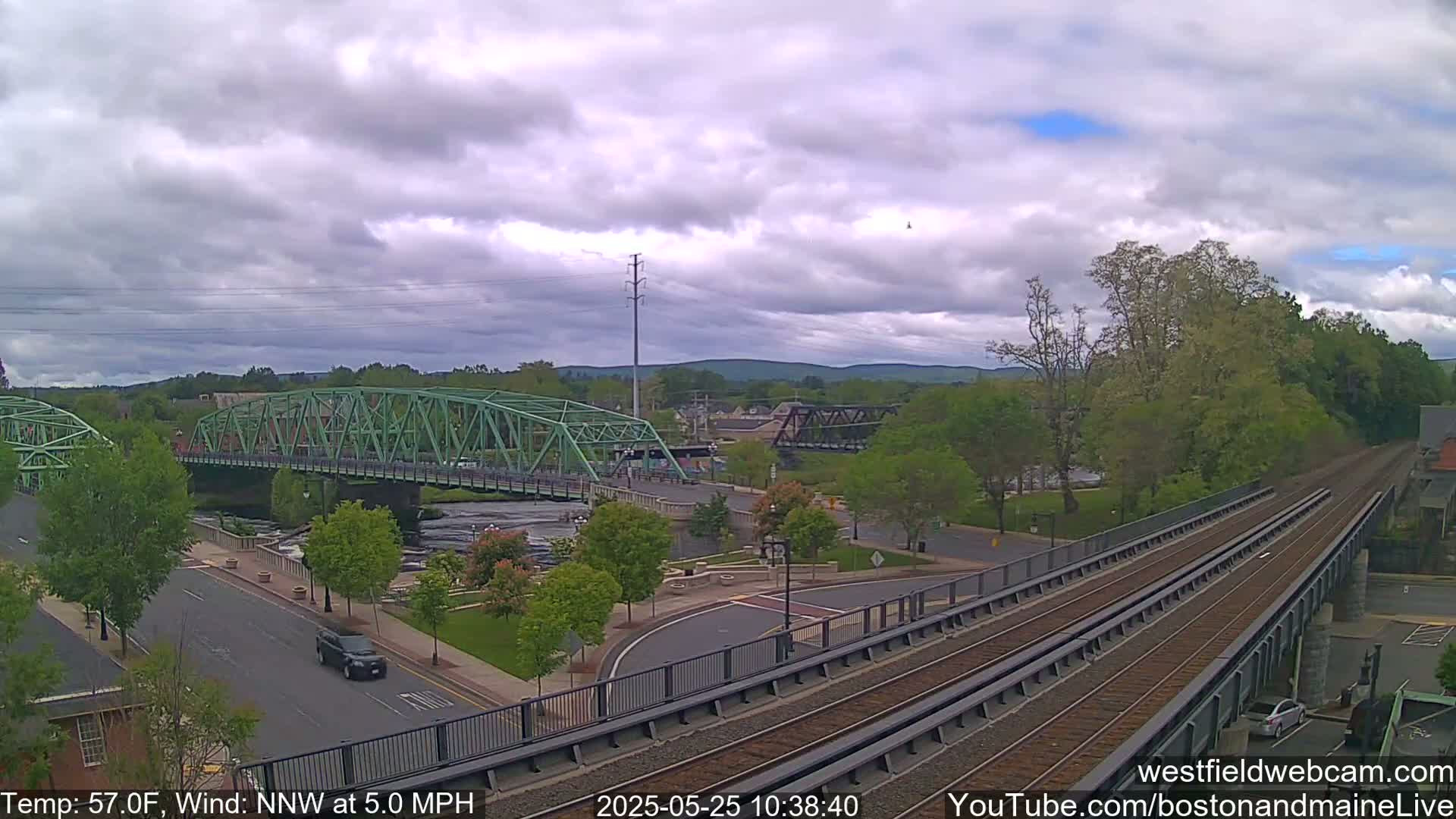 A partly cloudy day reveals a town scene with two bridges over a river, a road, train tracks, and lush green trees.