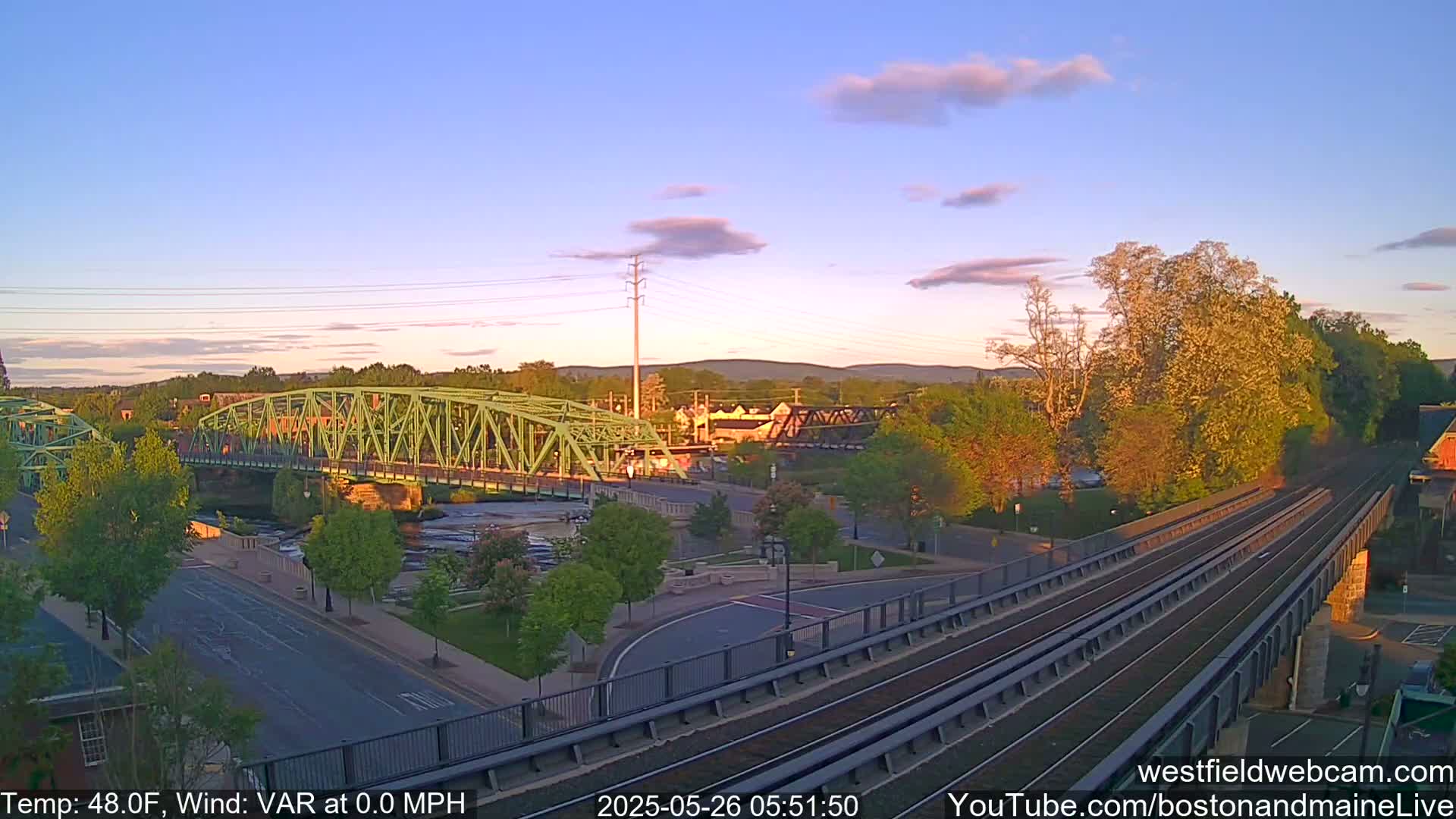 A green bridge spans a river, with train tracks and a town visible under a partly cloudy sunset sky.