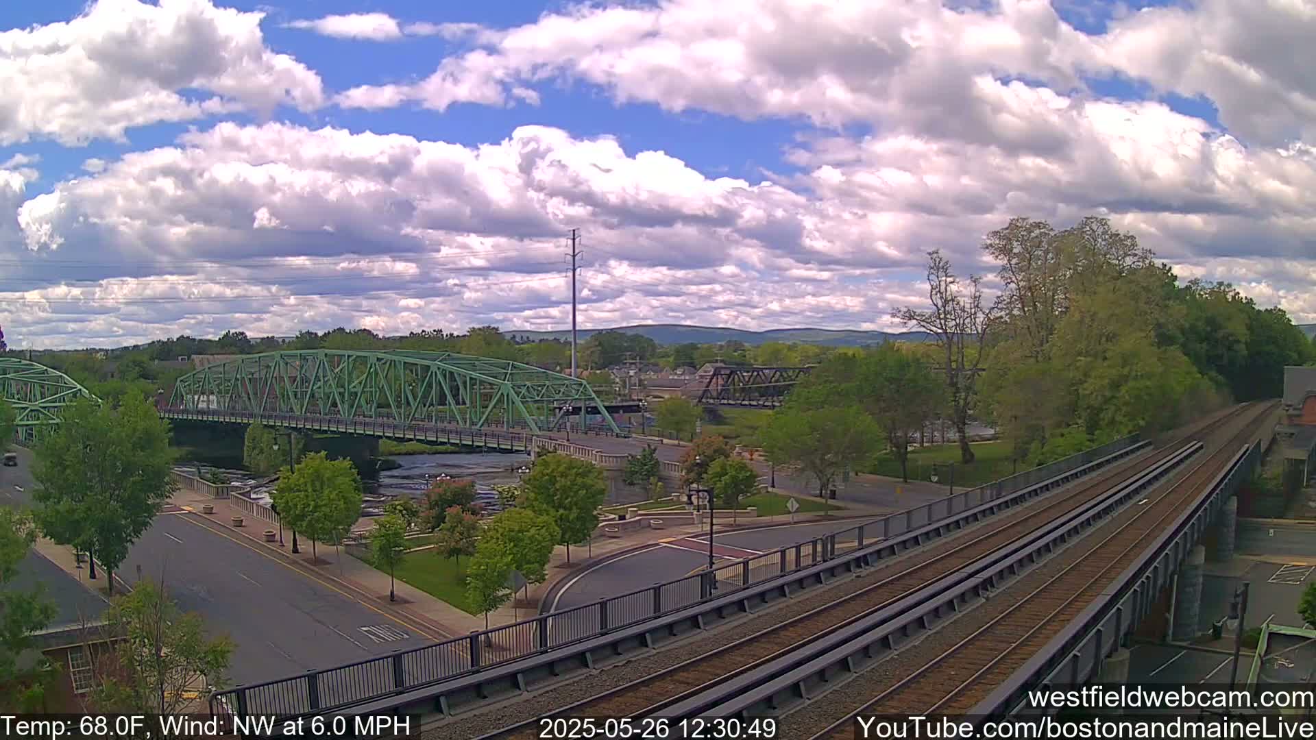 A partly cloudy day reveals a river scene with two bridges, one green and one dark metal, a roadway, and train tracks running alongside a park-like area with lush green trees.
