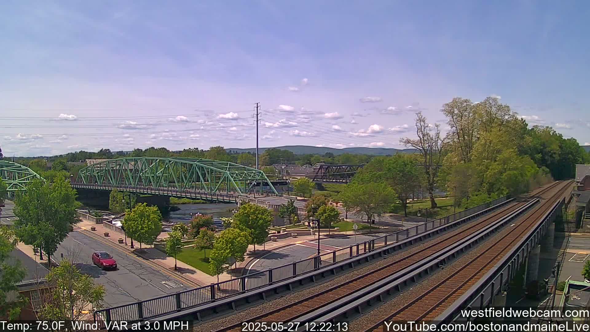 A view of a town with two bridges over a river, a railway track, and lush green trees under a partly cloudy sky.
