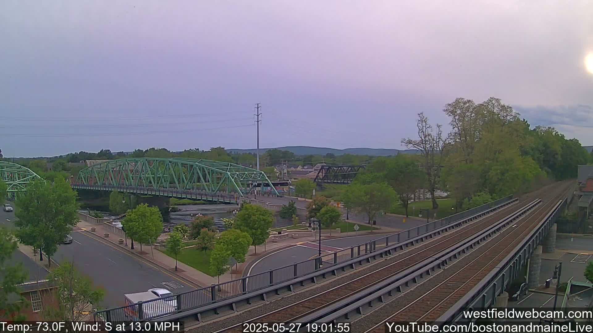 A green bridge spans a river in a town under a partly cloudy sky, with a train track elevated alongside a road.