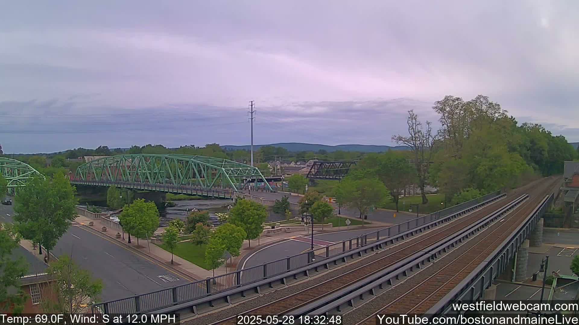 A view of a town with two bridges over a river, a train track overpass, and green trees under a partly cloudy sky.