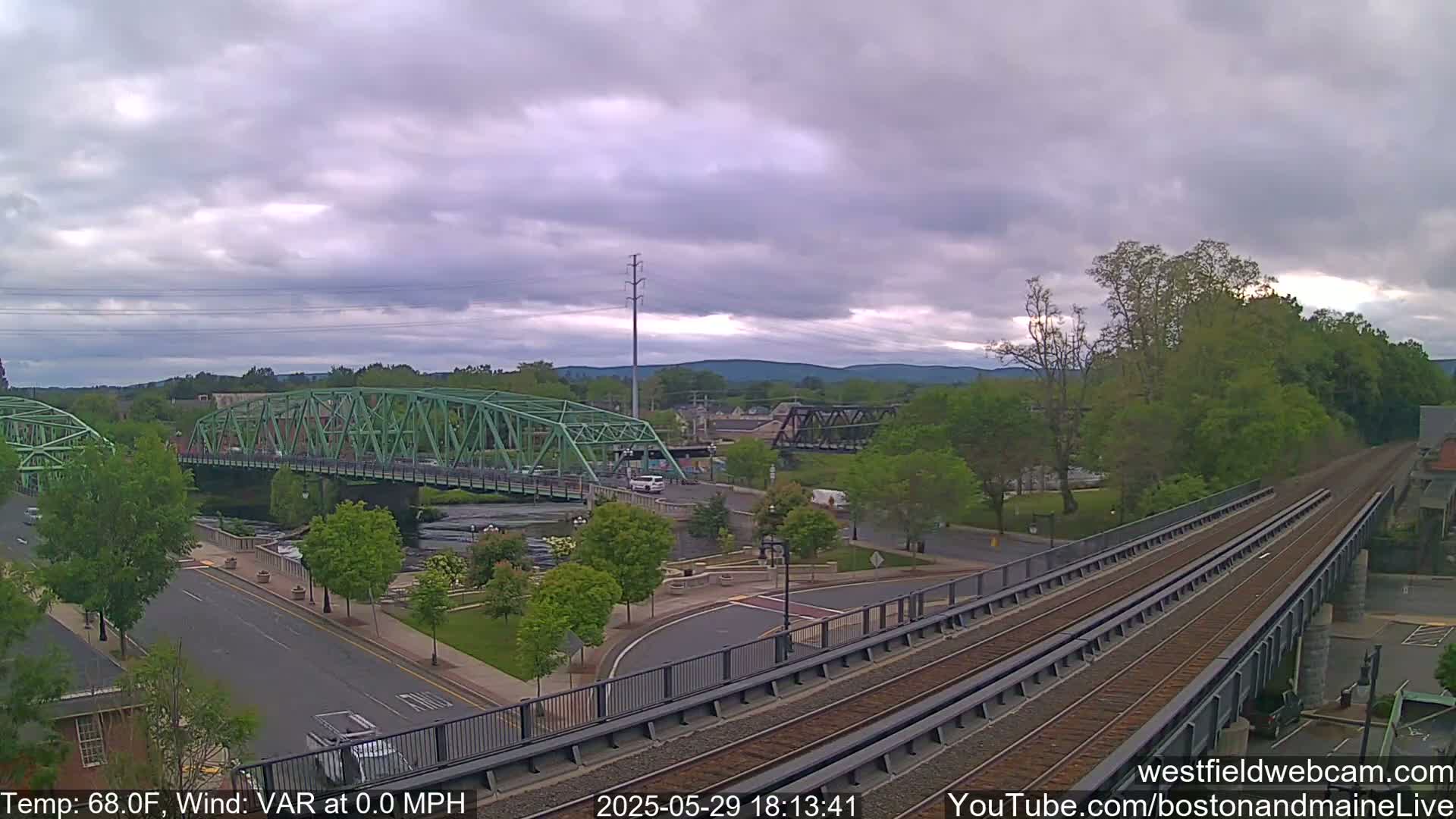 A view of a town under a cloudy sky shows two bridges over a river, with train tracks crossing in the foreground.