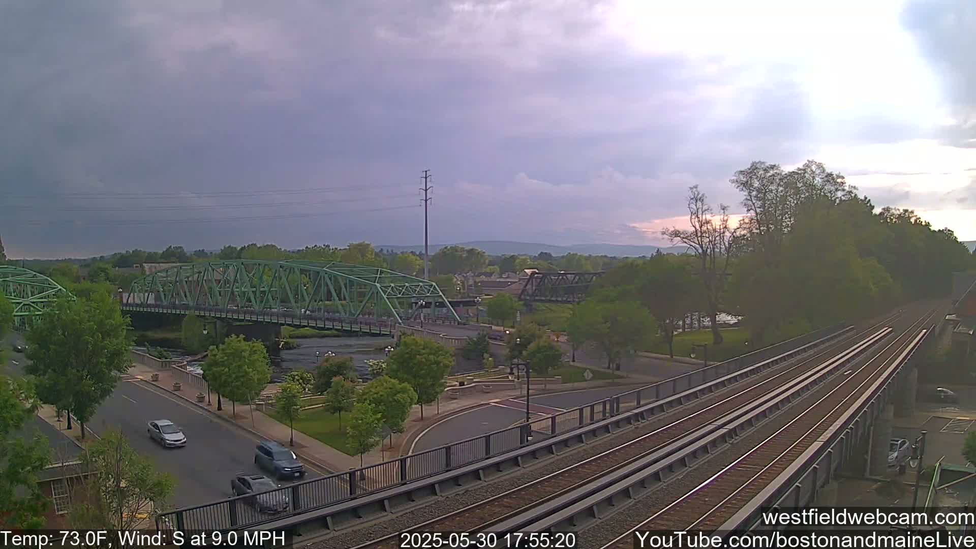 A green bridge spans a river in a town under partly cloudy skies, with train tracks visible in the foreground.