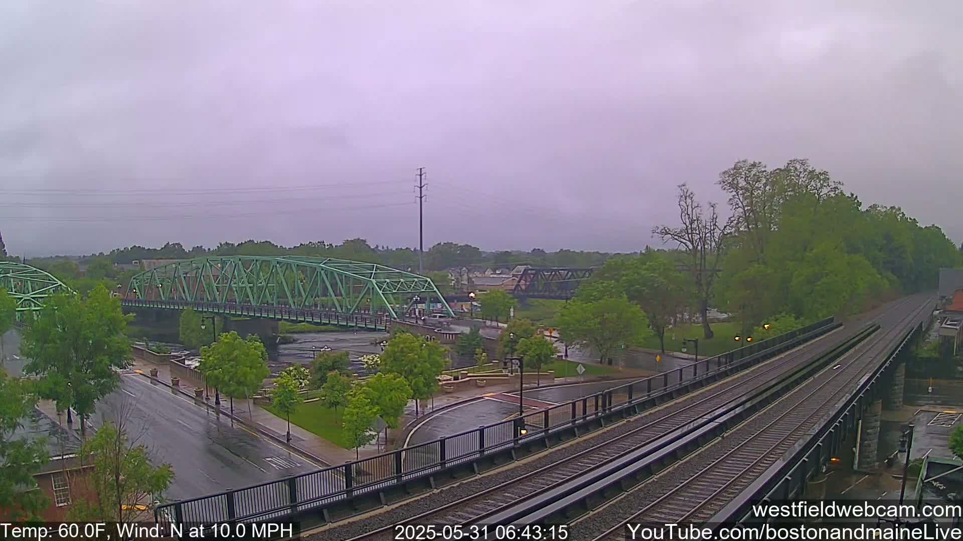 A view of a river, two bridges, and train tracks under an overcast sky, with rain falling on wet roads and streets.