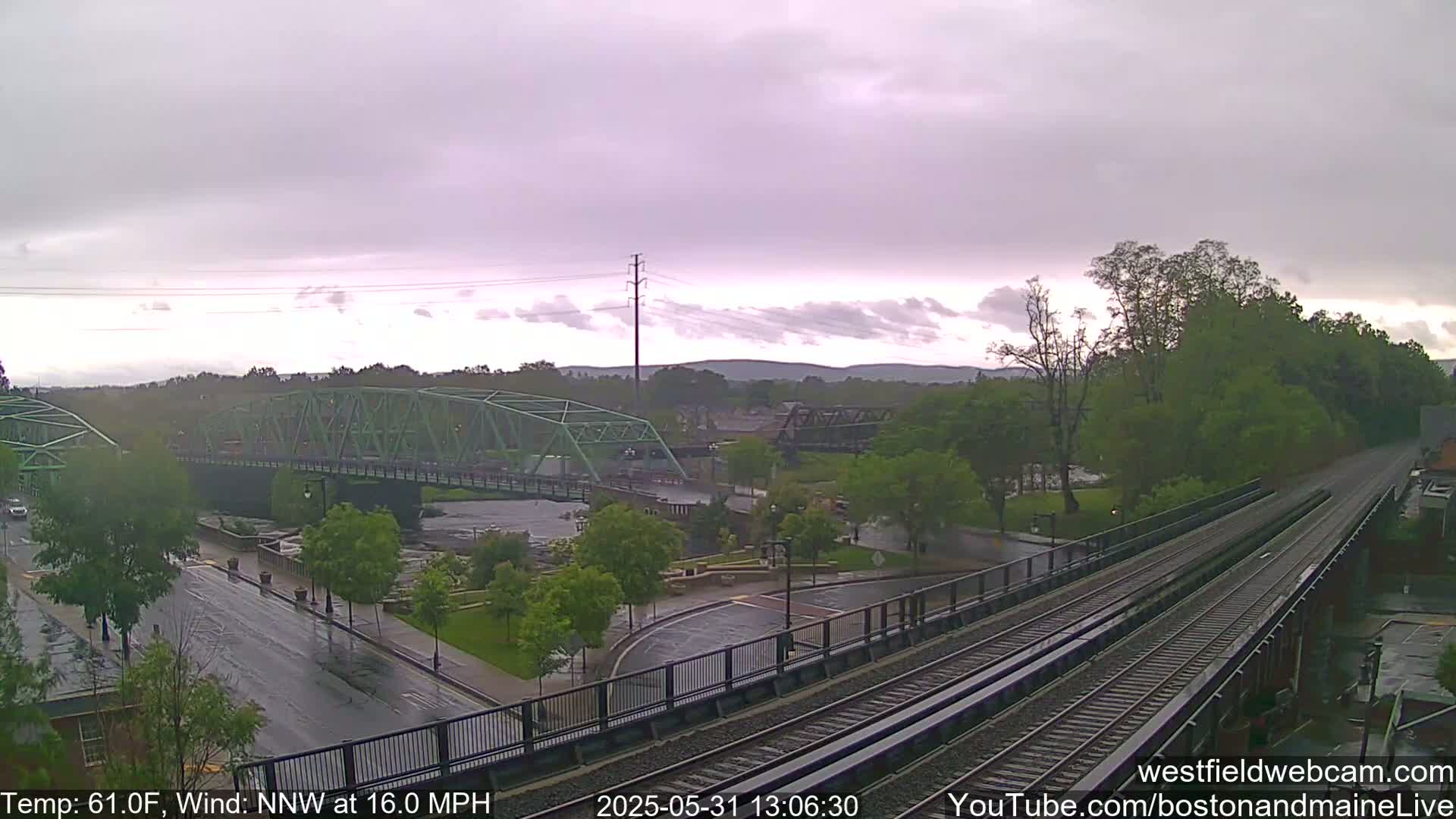 A view of a town under a cloudy sky, showing a green bridge over a river, a park, and train tracks.