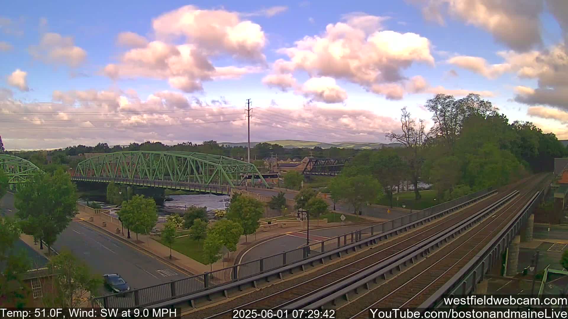 A green bridge spans a river beside a train track under a partly cloudy sky.