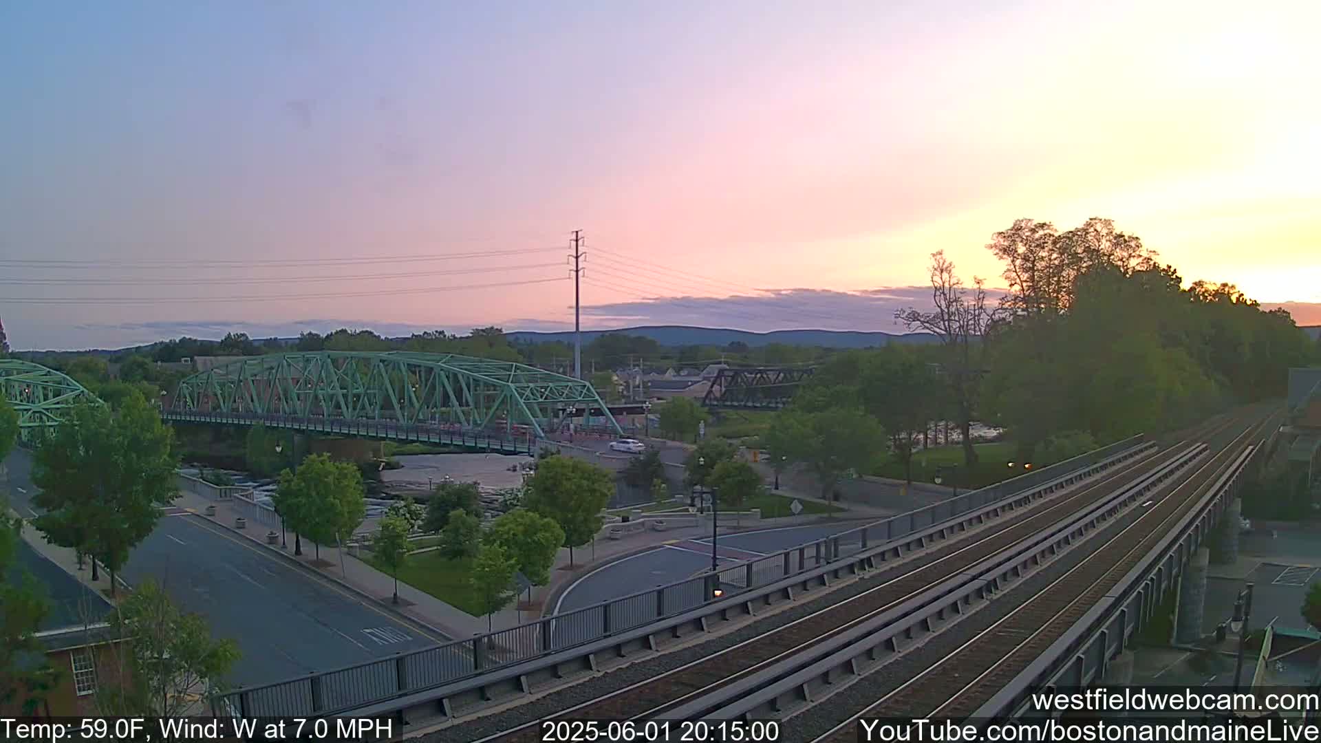 A pastel sunset illuminates a town scene featuring a green bridge over a river, train tracks, and surrounding trees and buildings.
