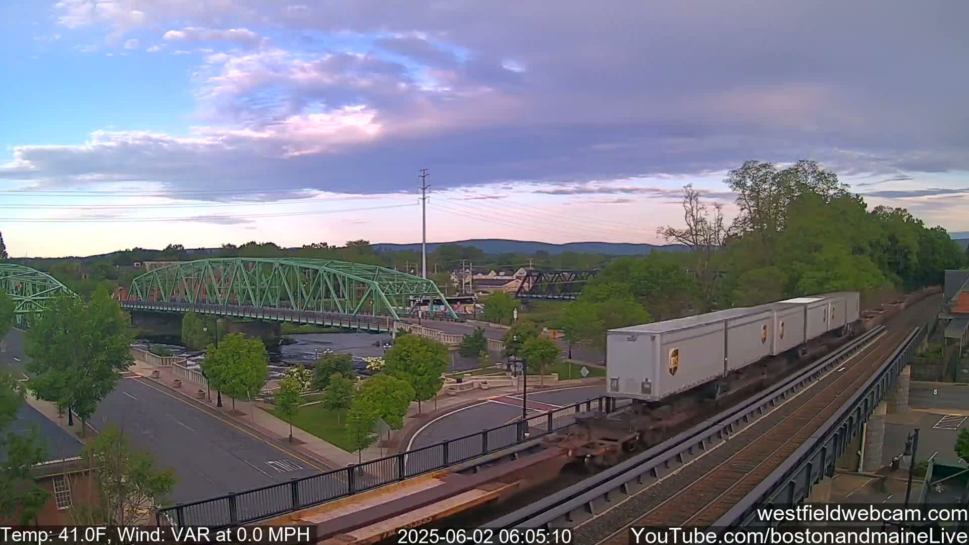 A train carrying several large cargo containers passes over a bridge, with a smaller green bridge visible in the background under a partly cloudy sky, the temperature is 41°F, and the wind is variable at 0.0 mph.