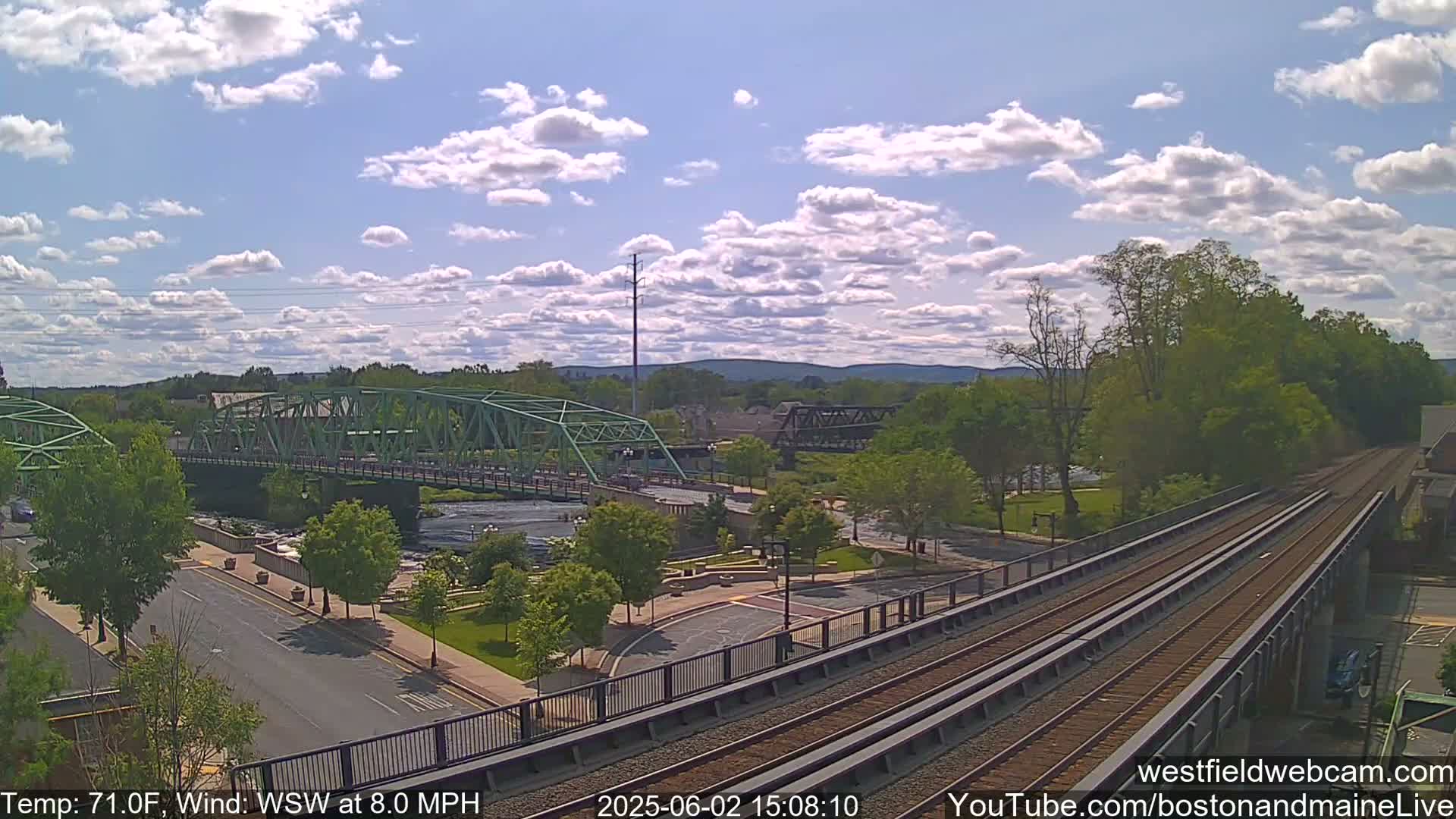 A partly cloudy day shows a view of two bridges, a river, a roadway, train tracks, and lush green trees.