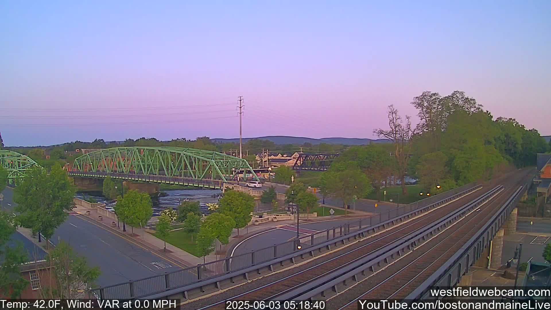 A view of a river with two bridges, one green and one dark metal, under a pale pink twilight sky.