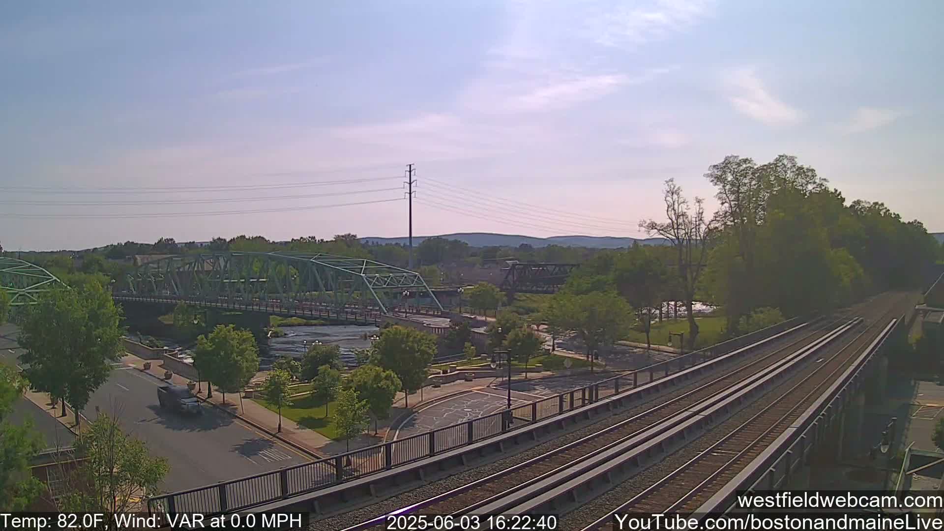 A view from a train trestle on a sunny day shows a river, two bridges, a road, and a park below.