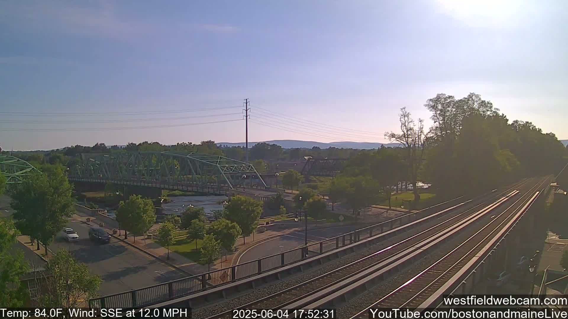 A high-angle view of a town on a sunny day with a light wind shows a green bridge over a river, a railroad track, and cars driving on a road, all surrounded by trees.