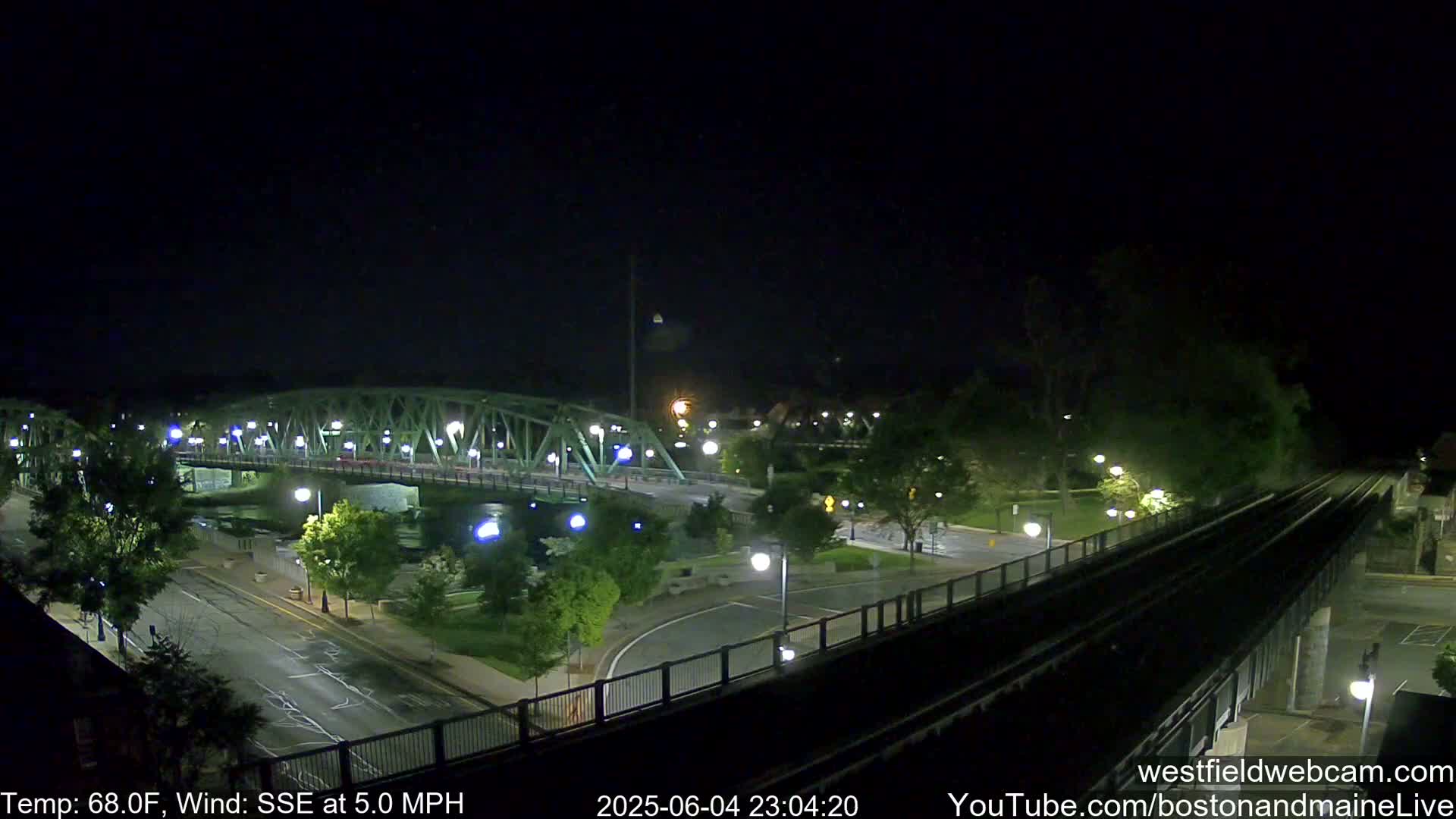 A nighttime view of a bridge over a waterway, with a roadway and train tracks visible, under a clear, dark sky and a 5 mph wind from the south-southeast.