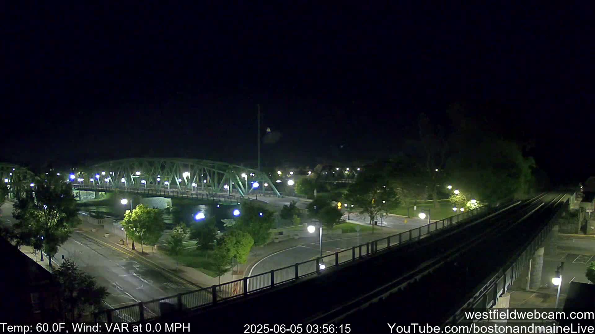A nighttime view of a green metal bridge over a river, with roads, trees, and train tracks visible under a clear, dark sky.