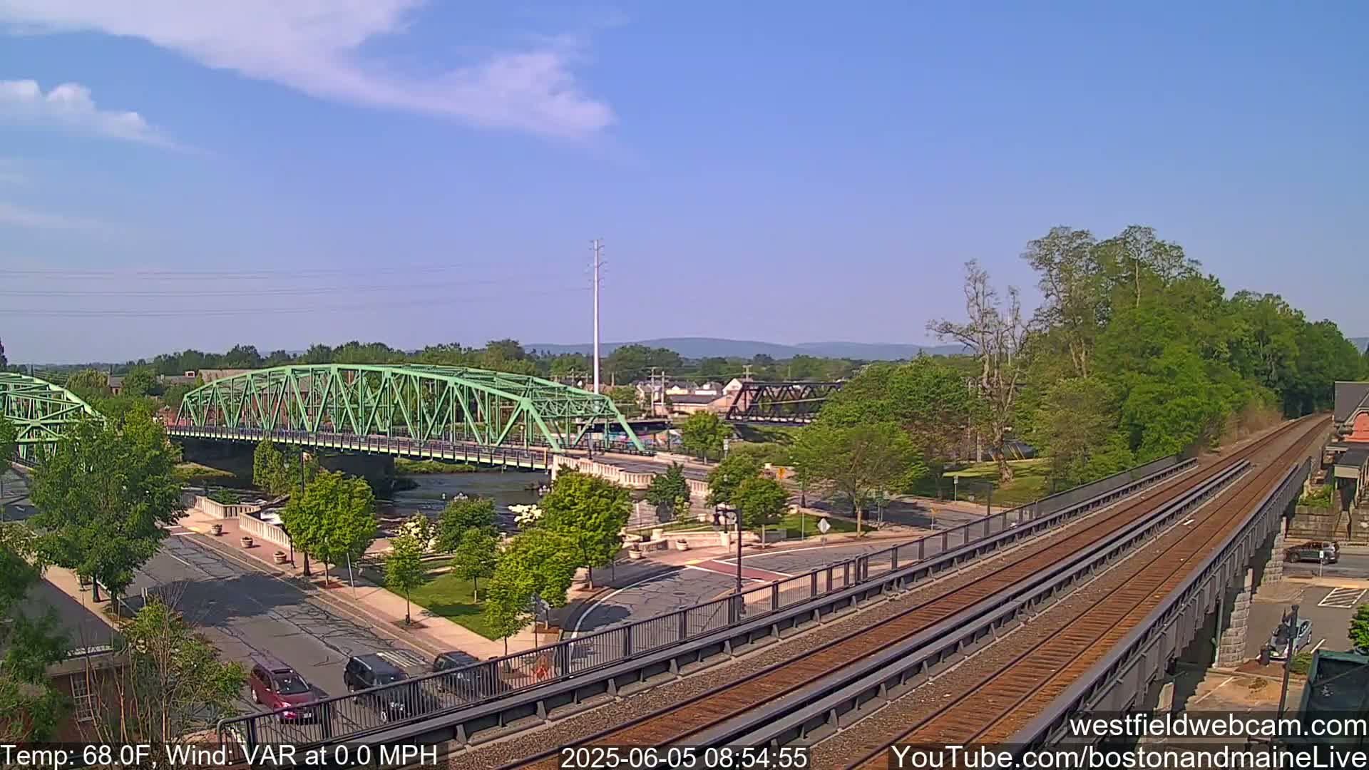 A sunny day shows a view of a river with two bridges, one green and one dark metal, train tracks crossing on a high bridge, and a town in the background.