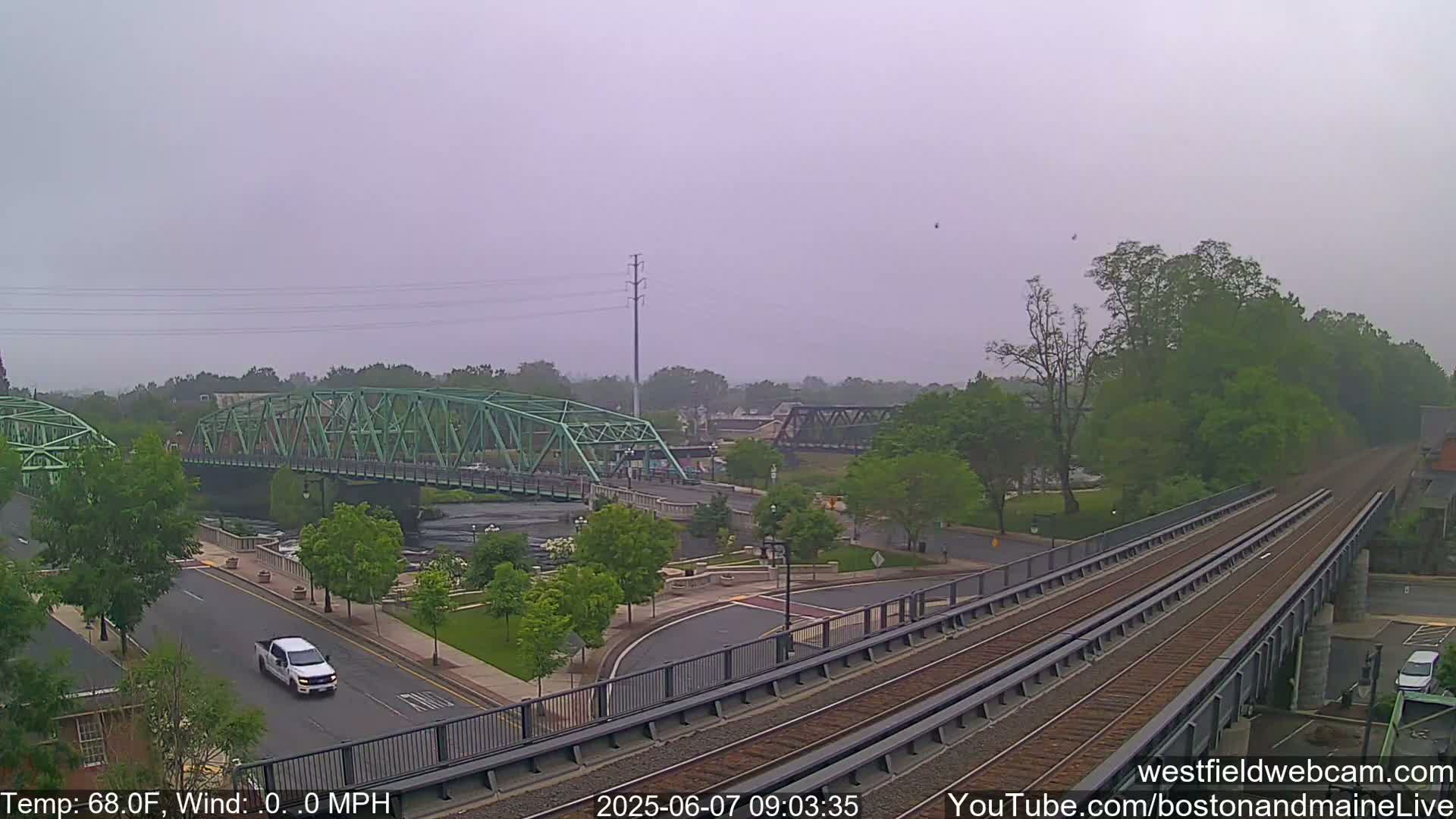 A view of a town on an overcast day, featuring two bridges over a river, a roadway with a white truck, and train tracks crossing in the foreground.