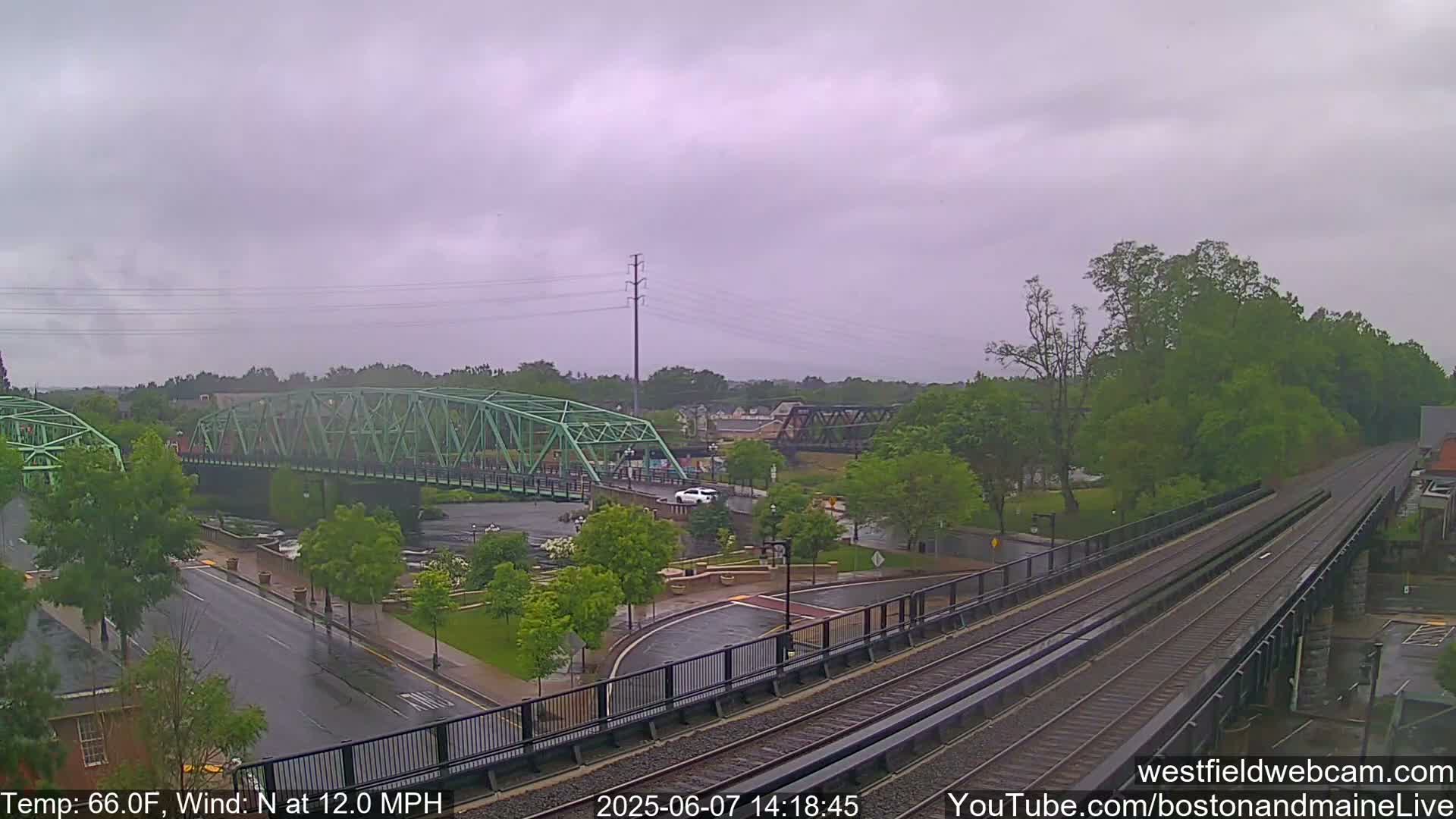 A view of a wet town with two bridges over a river, train tracks, and lush green trees under an overcast sky.