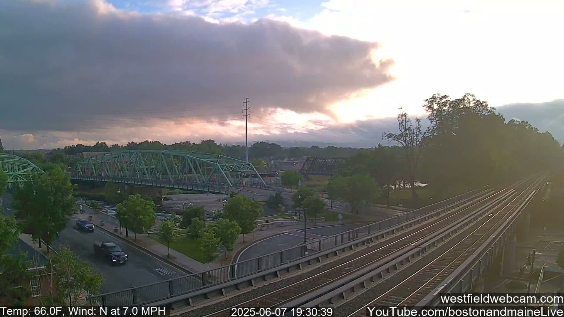 A partly cloudy sunset illuminates a river scene with a green metal bridge, a roadway, and train tracks, with a temperature of 66°F and a north wind at 7 mph.