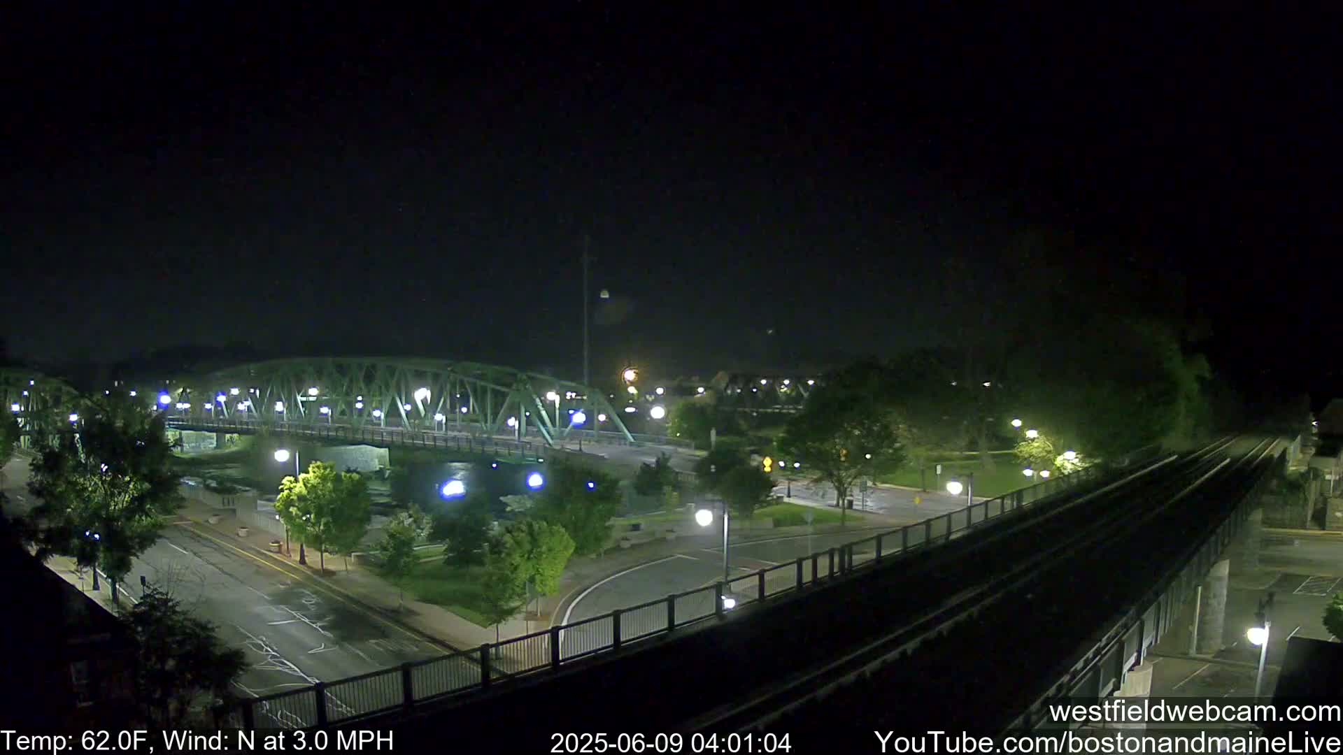 A nighttime view shows a bridge over a waterway, surrounded by roads, trees, and streetlights, with a clear sky and a light north wind.